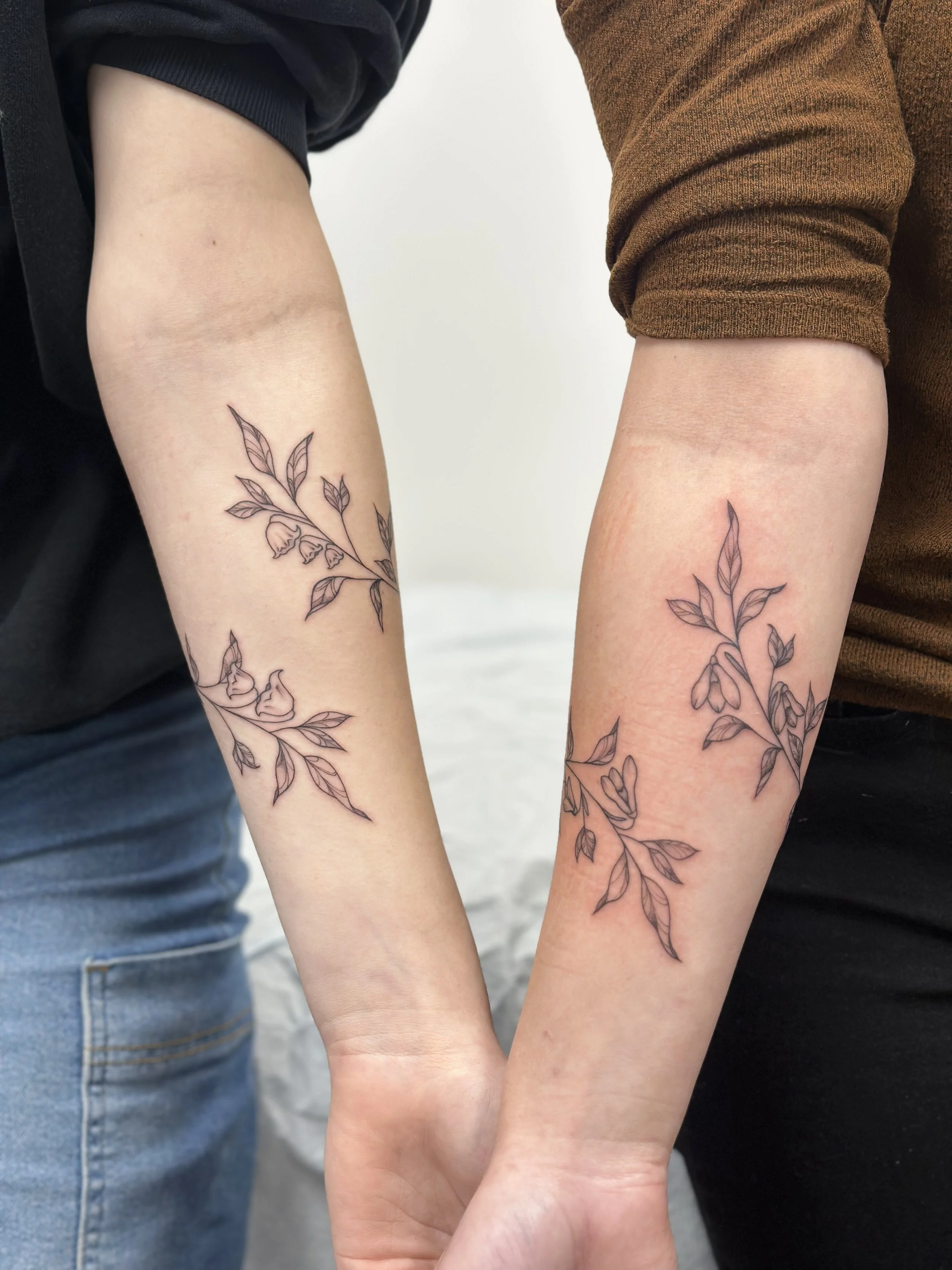 Two people with matching black ink floral tattoos on their forearms, holding hands with intertwined fingers.