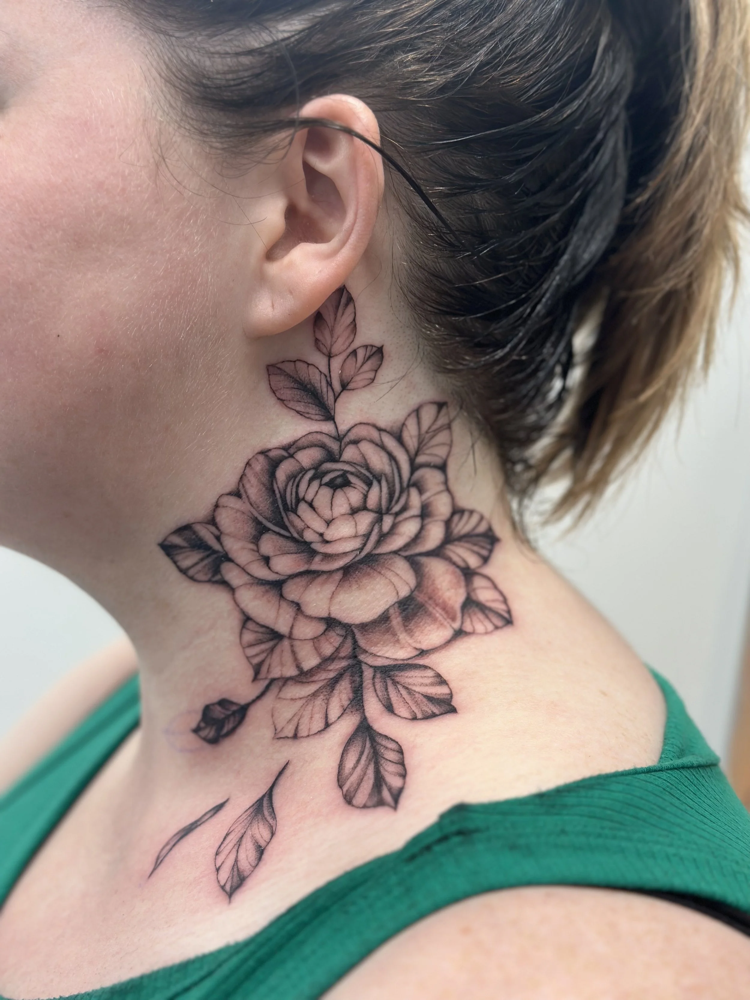 A close-up of a person's neck tattoo featuring a detailed black and gray blooming rose with leaves.