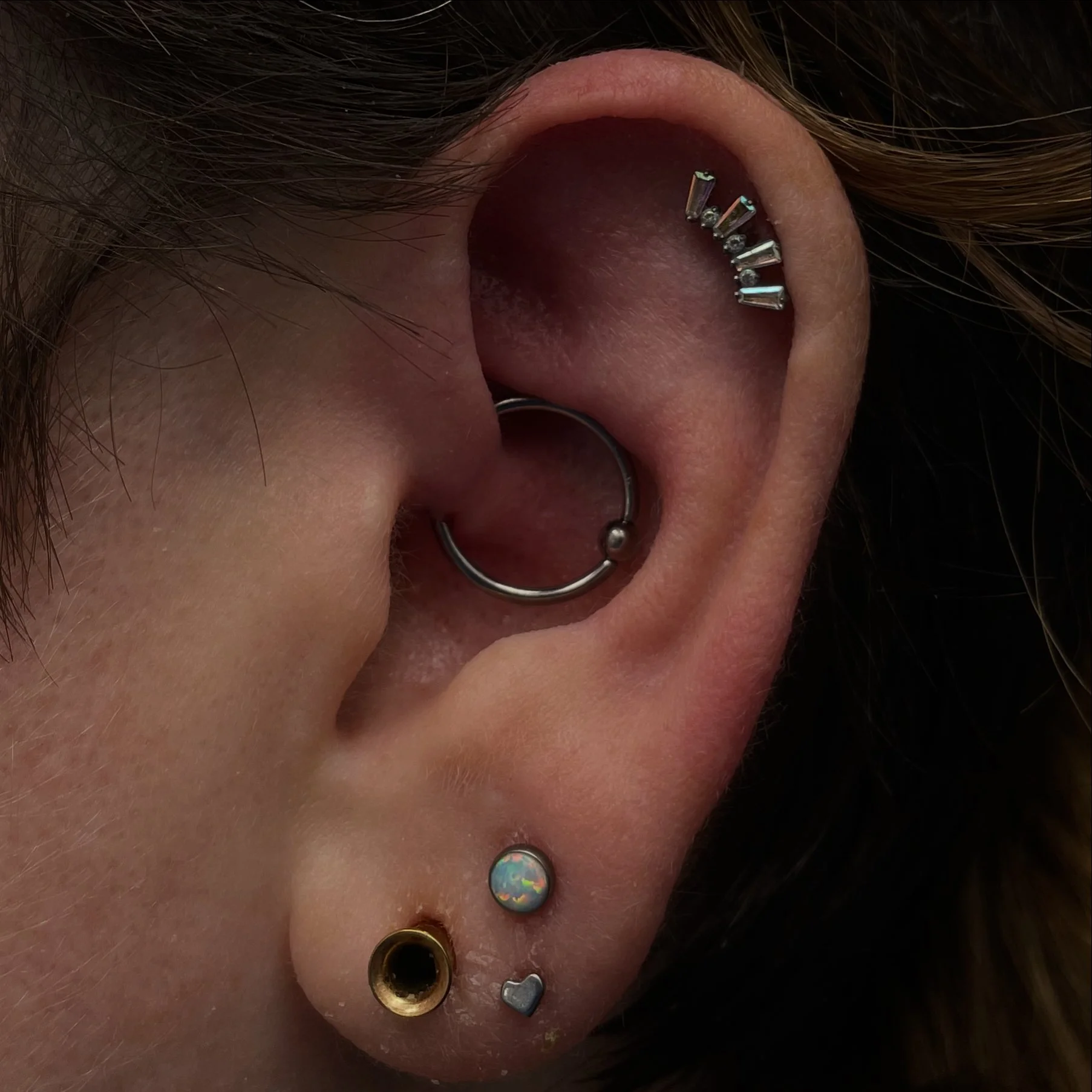 Close-up of a person's multiple ear piercings, including a cartilage pierce with metallic barbell jewelry, a helix piercing with multiple stud earrings, a tragus piercing, a helix piercing with an opal stud, a lobe piercing with a gold ring, and a lo