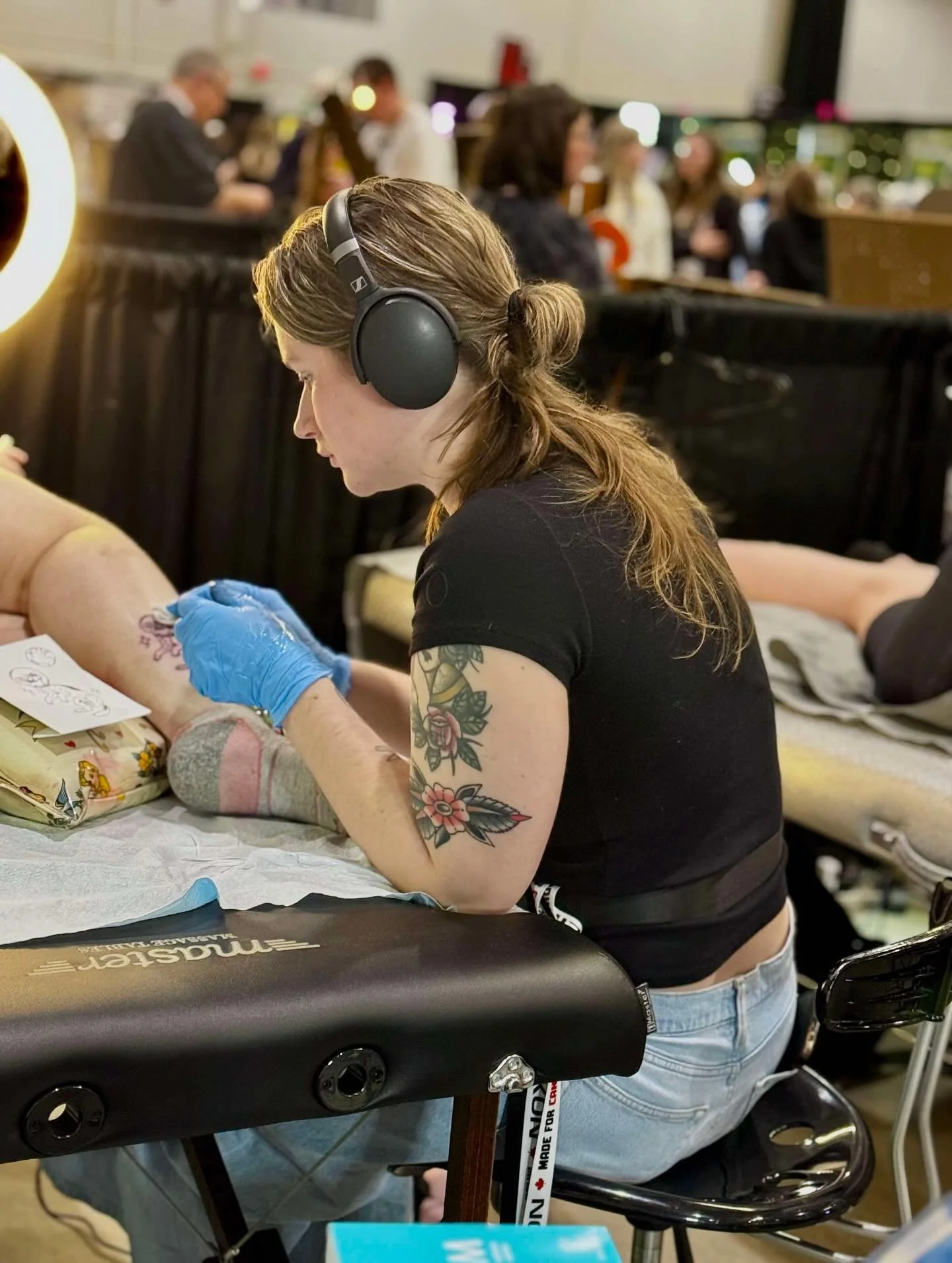 Tattoo artist with long hair, wearing a black shirt and blue gloves, doing a tattoo on a person's leg in a busy tattoo studio, while wearing headphones.
