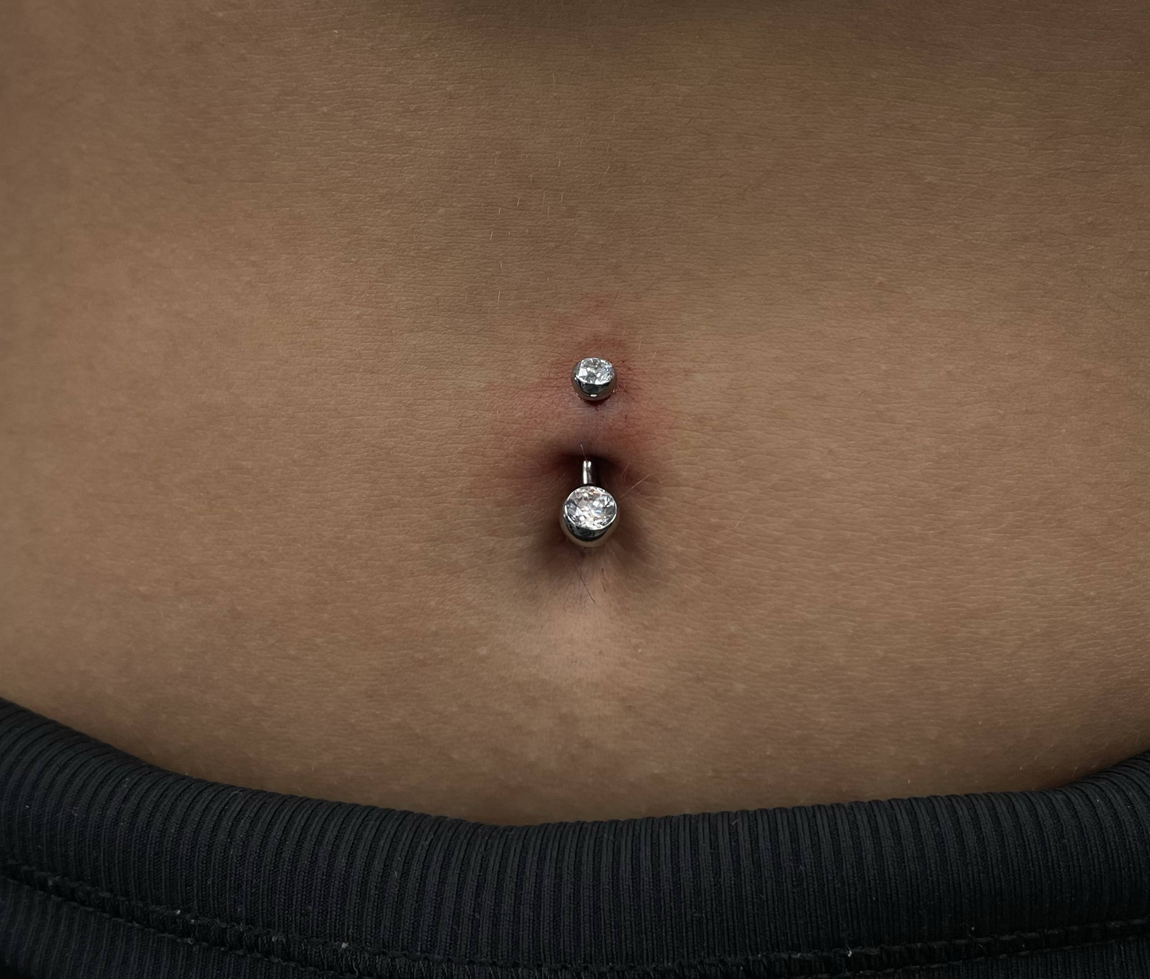 Close-up of a person's navel with a belly button piercing featuring a curved barbell with two clear gemstones.