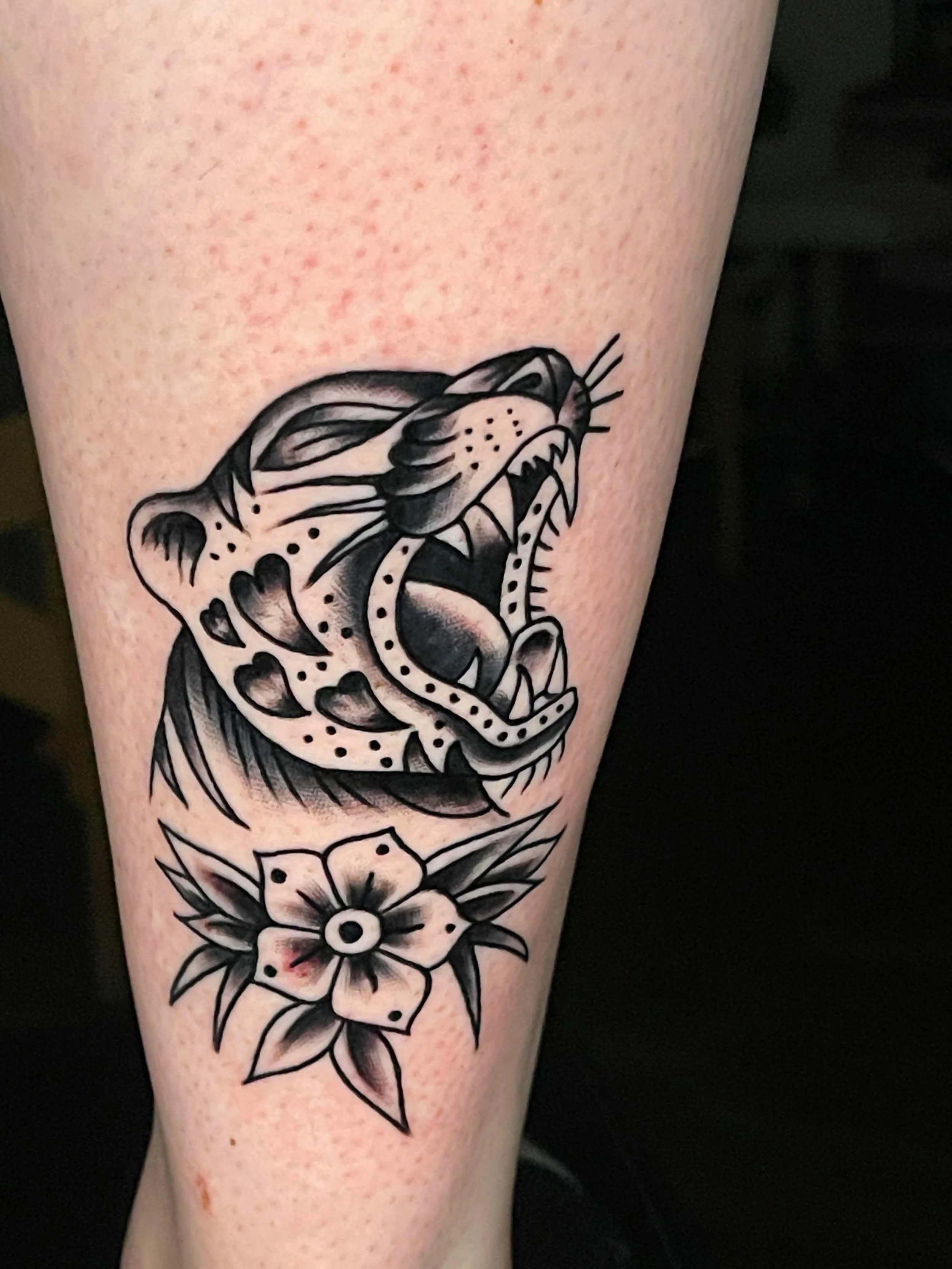 A black and grey tattoo of a roaring big cat's head with a flower below it on a person's arm.