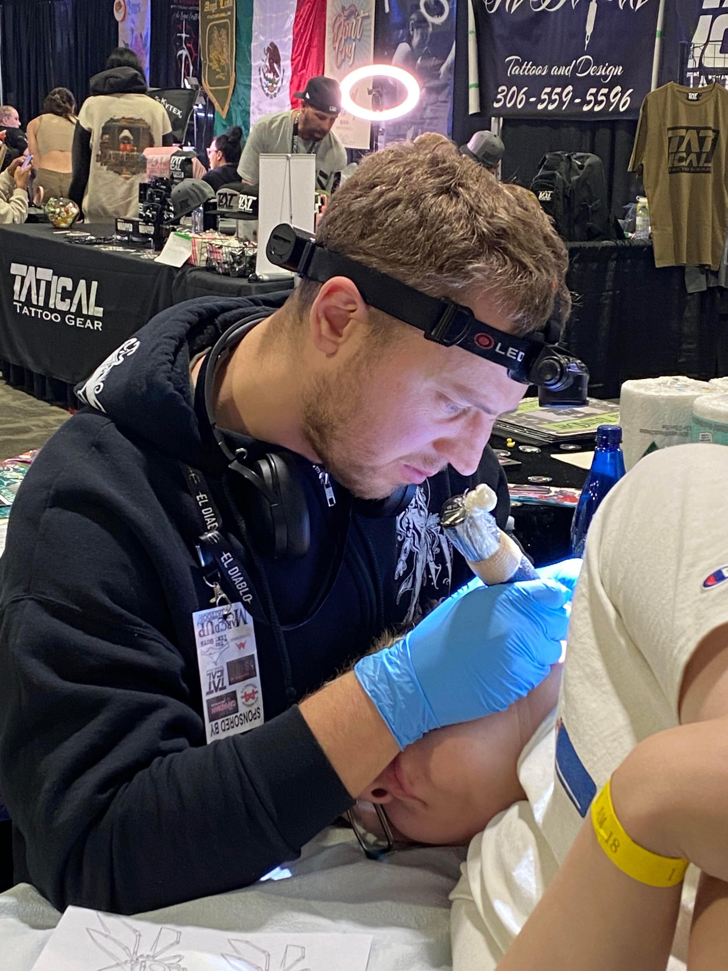 A tattoo artist wearing a headlamp and blue gloves tattooing a person's arm at a convention, with booths and banners in the background.