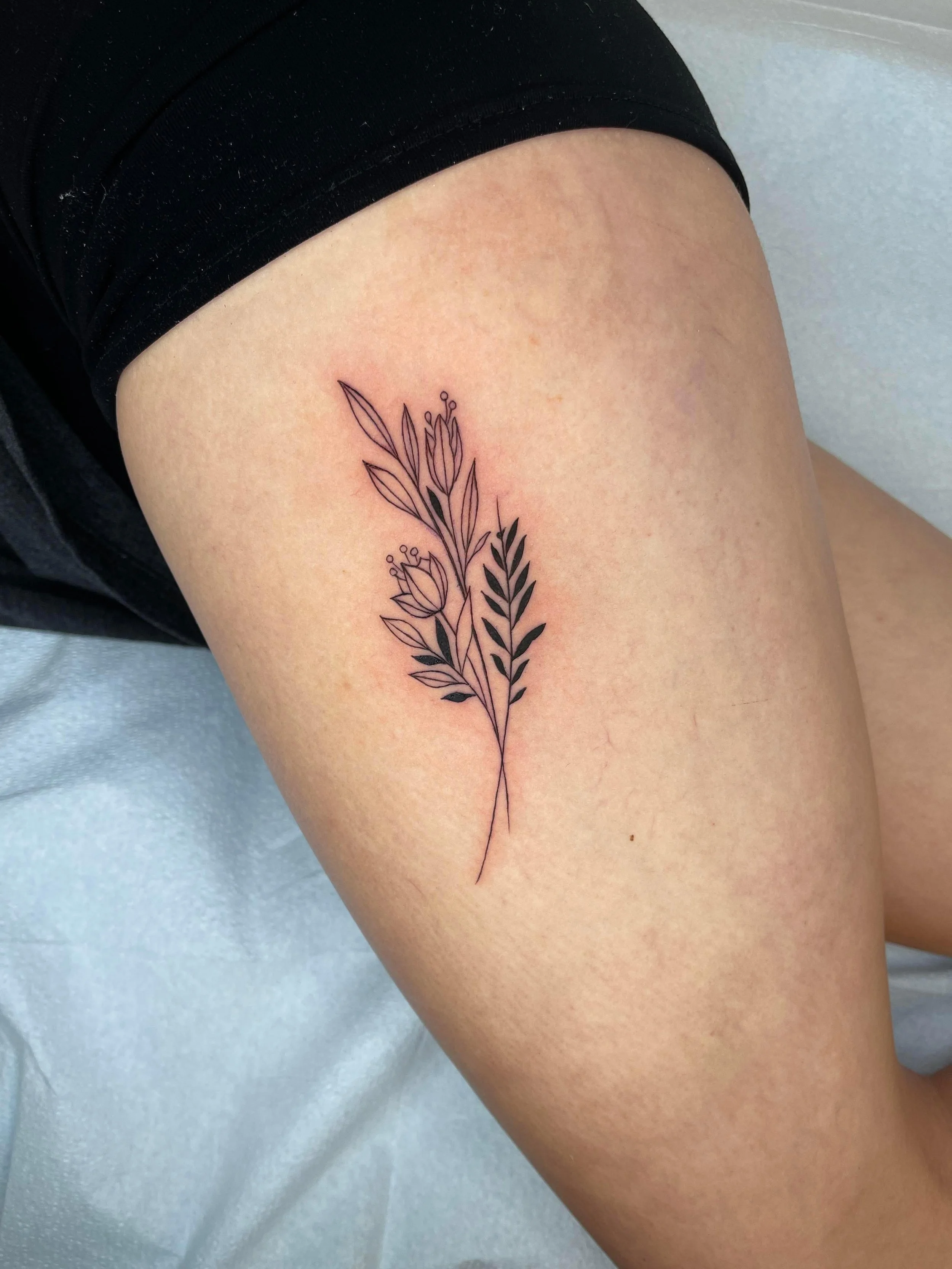 A tattoo of a simple black line drawing of a branch with leaves and flowers on a person's thigh.