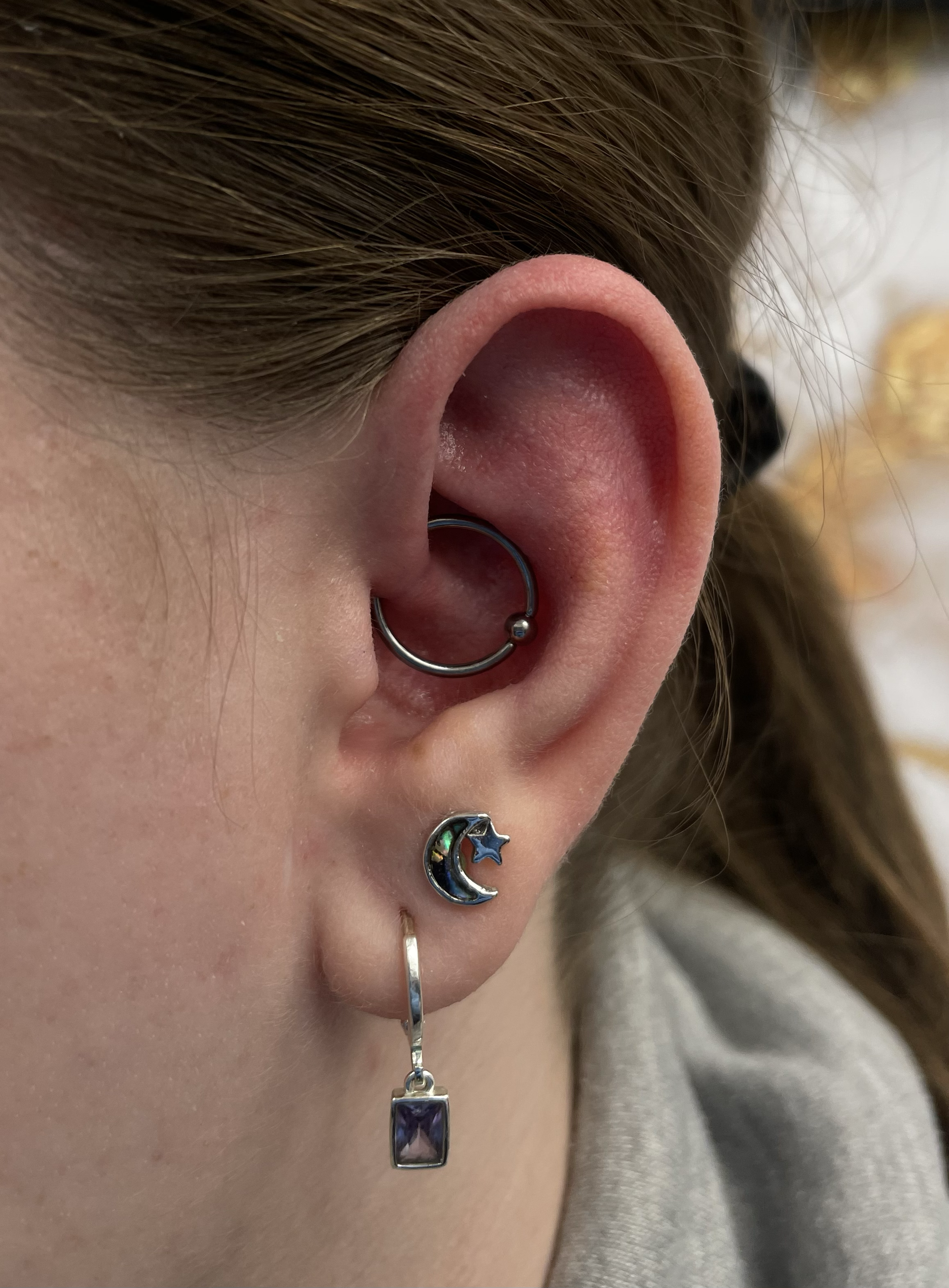 Close-up of a person's ear with three earrings: a silver hoop in the helix, a celestial-themed stud earring with a moon and star in the lobe, and a dangling earring with a purple gemstone.