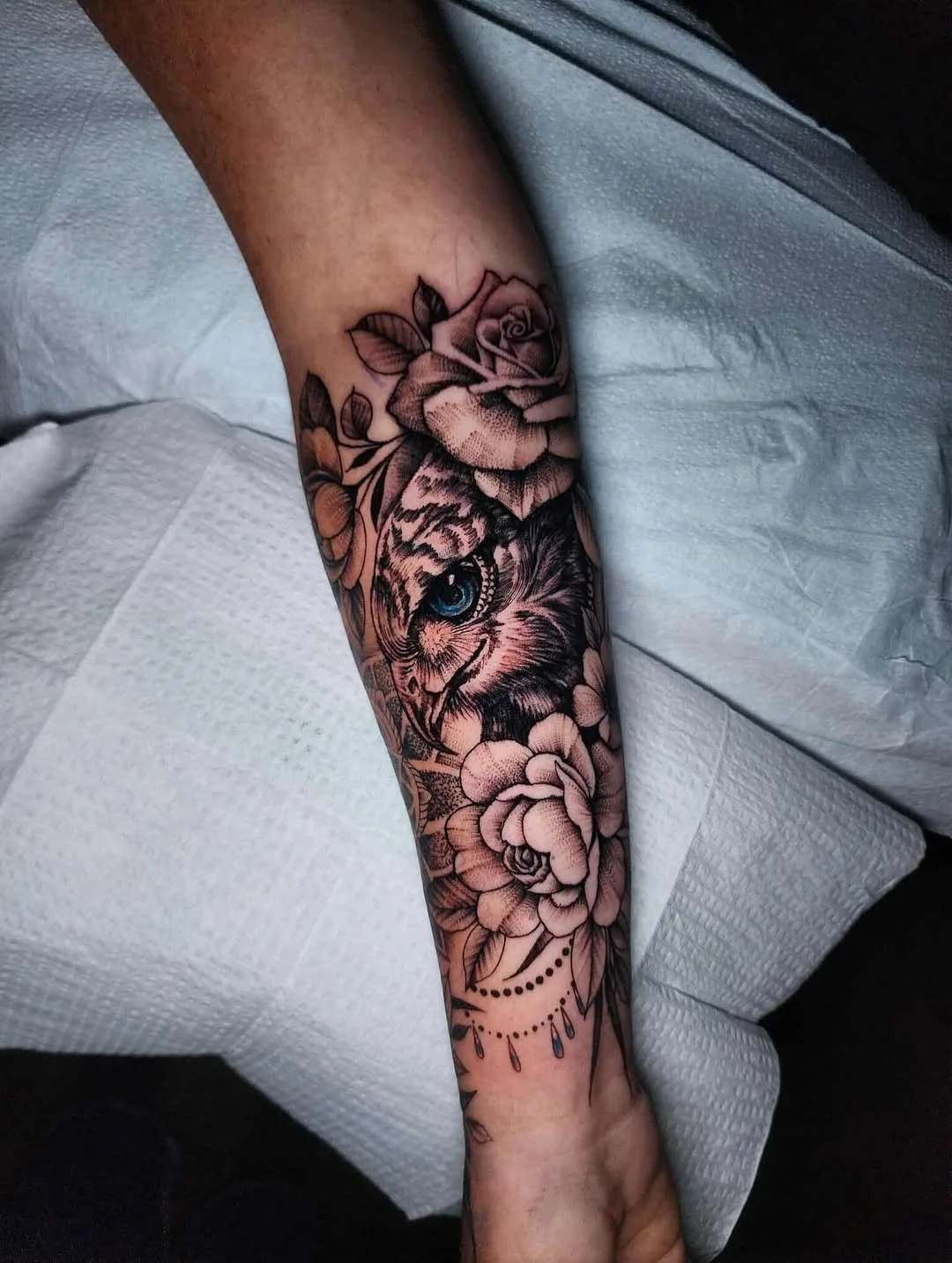 Tattoo of a lion's face with an eye pattern, surrounded by roses and floral designs, on a person's forearm.