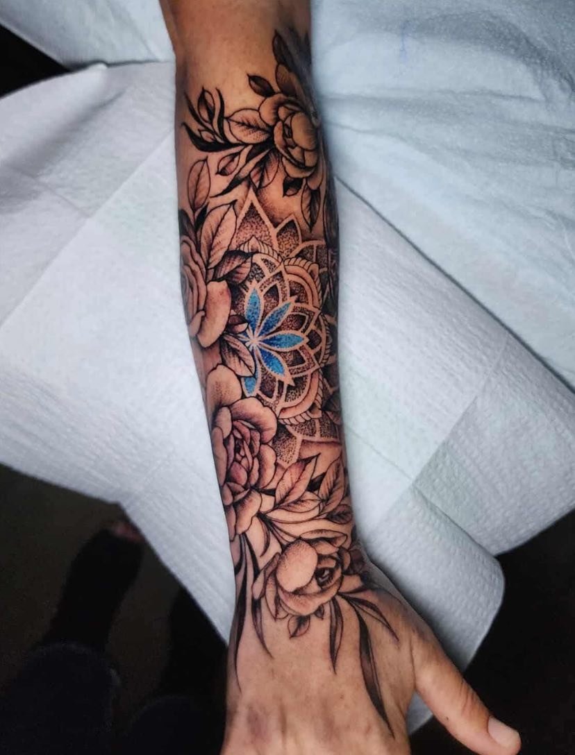 A detailed black and gray floral tattoo on a person's forearm and wrist, featuring roses, leaves, and a mandala design with blue accents.