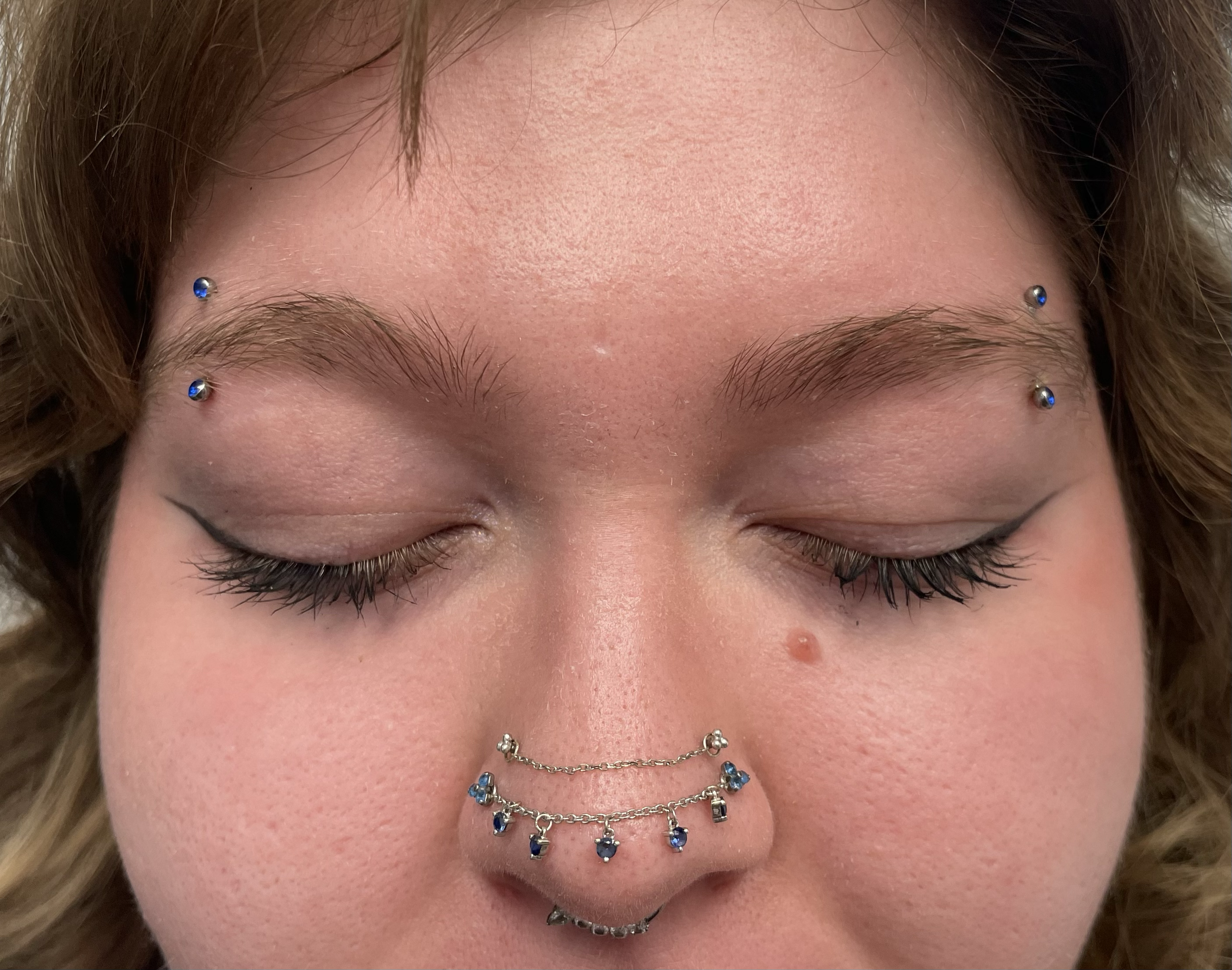 Close-up of a person's face with closed eyes, showing multiple facial piercings, including blue gemstone studs on eyebrows and a chain with charms on the nose.