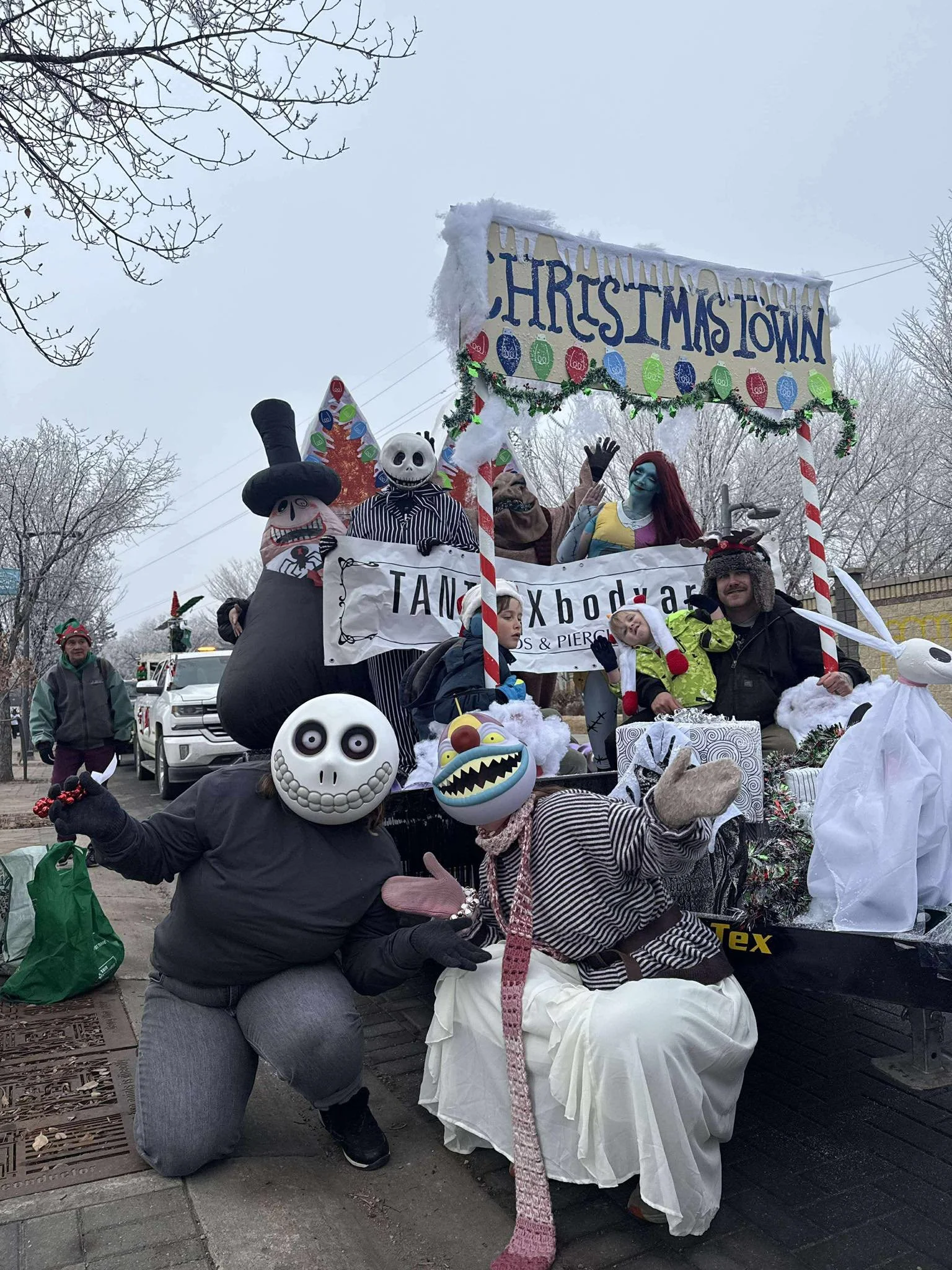 People dressed in Halloween costumes on a decorated parade float for Christmas Town, with masks of Jack Skellington, Sally, and other characters from The Nightmare Before Christmas, and a sign reading 'Christmas Town'.