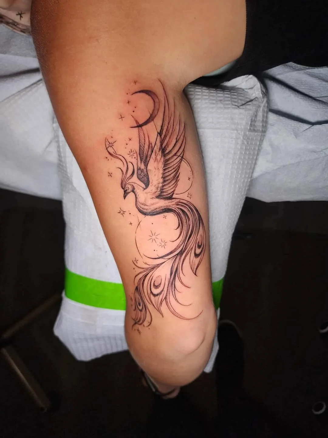 Tattoo of a stylized bird with spread wings and flowing tail feathers on a person's upper thigh, with a crescent moon and small stars surrounding the bird.