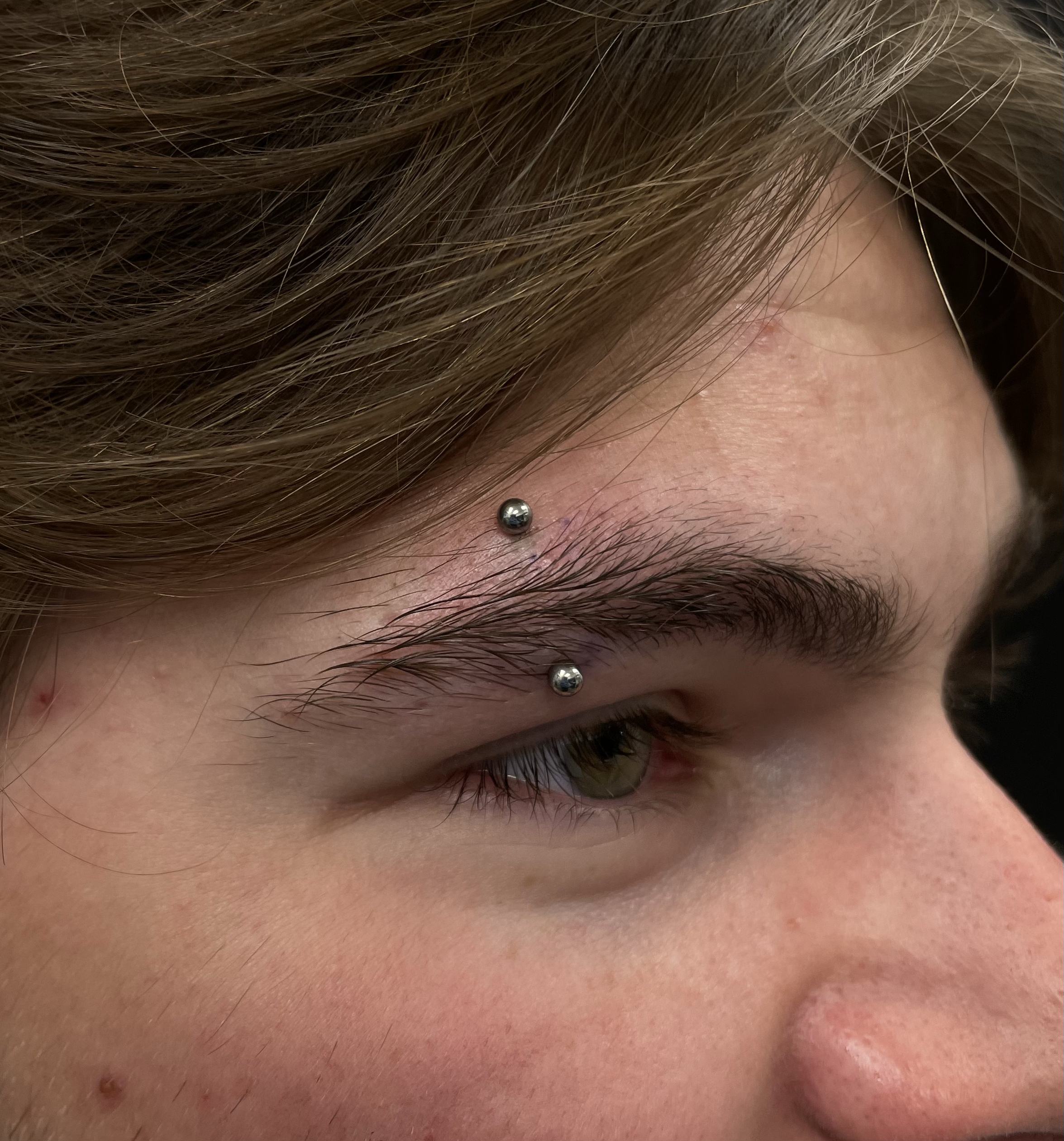 Close-up of a person's face showing their forehead, eyebrow with two dermal anchor piercings, and part of the eye and nose.