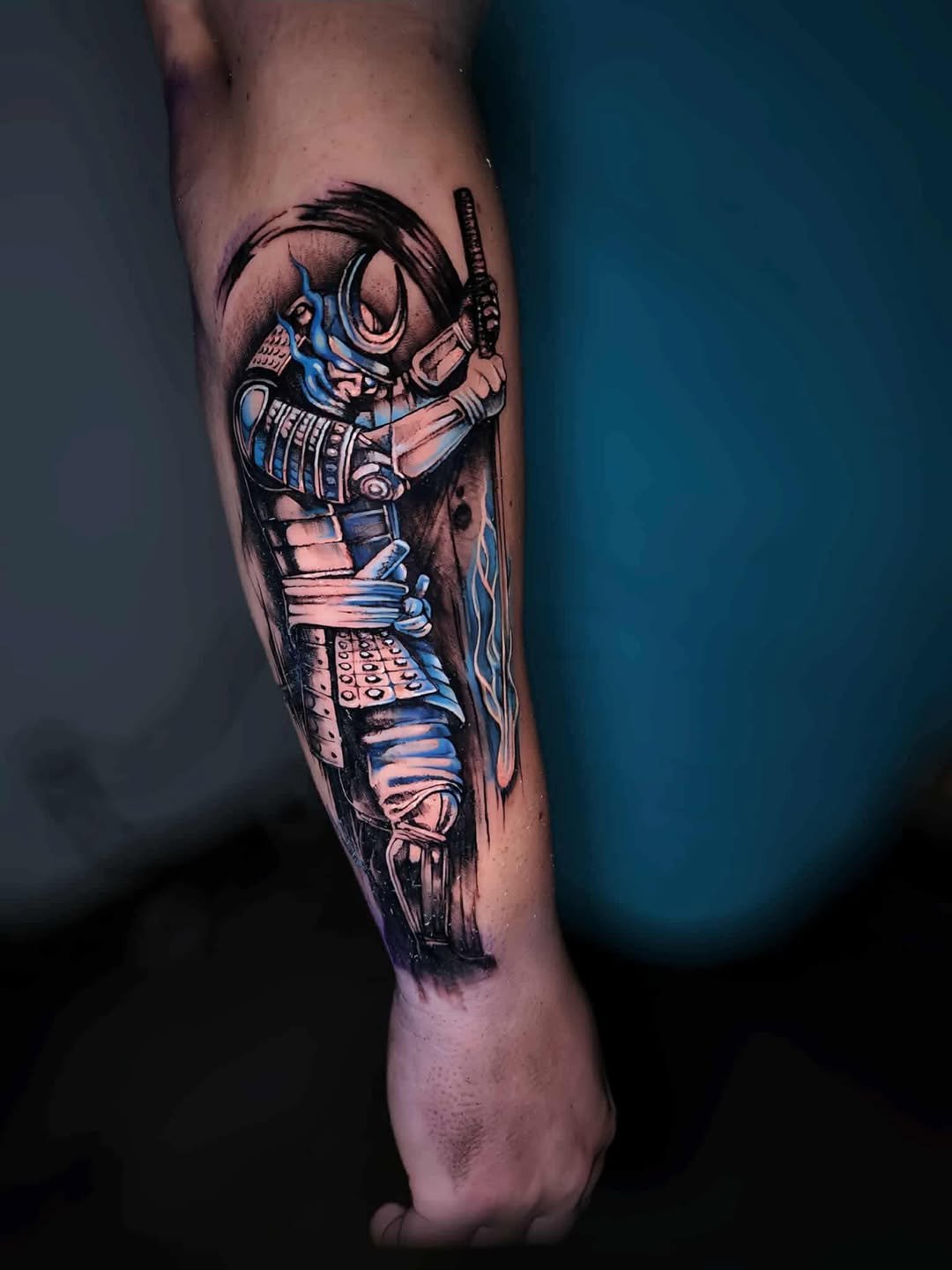Tattoo of a warrior woman in armor, holding a sword with electric blue accents, surrounded by a circular design, on a person's arm.