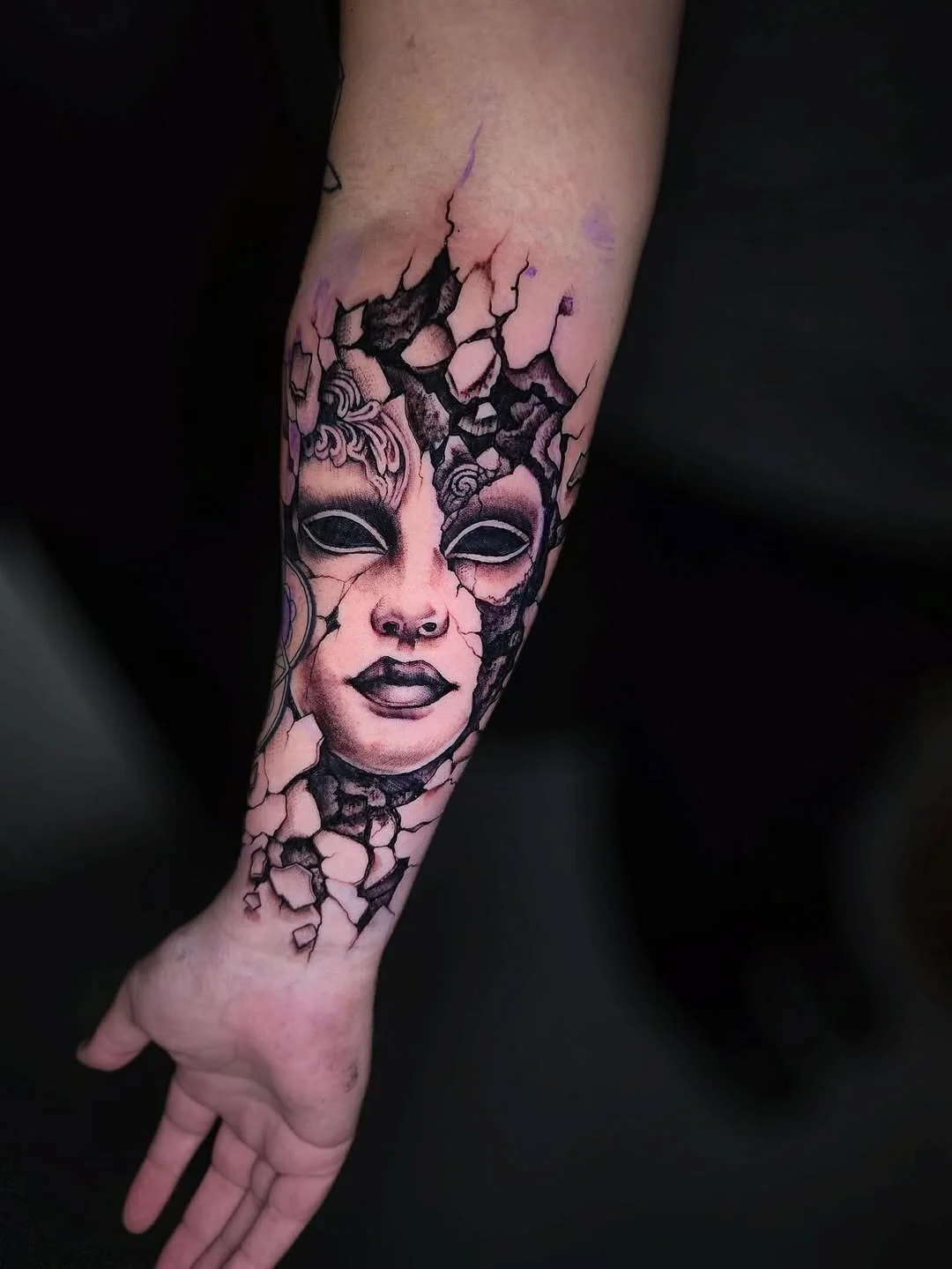 Tattoo of a woman's face with cracked surfaces, dark makeup, and empty black eyes on a person's forearm.