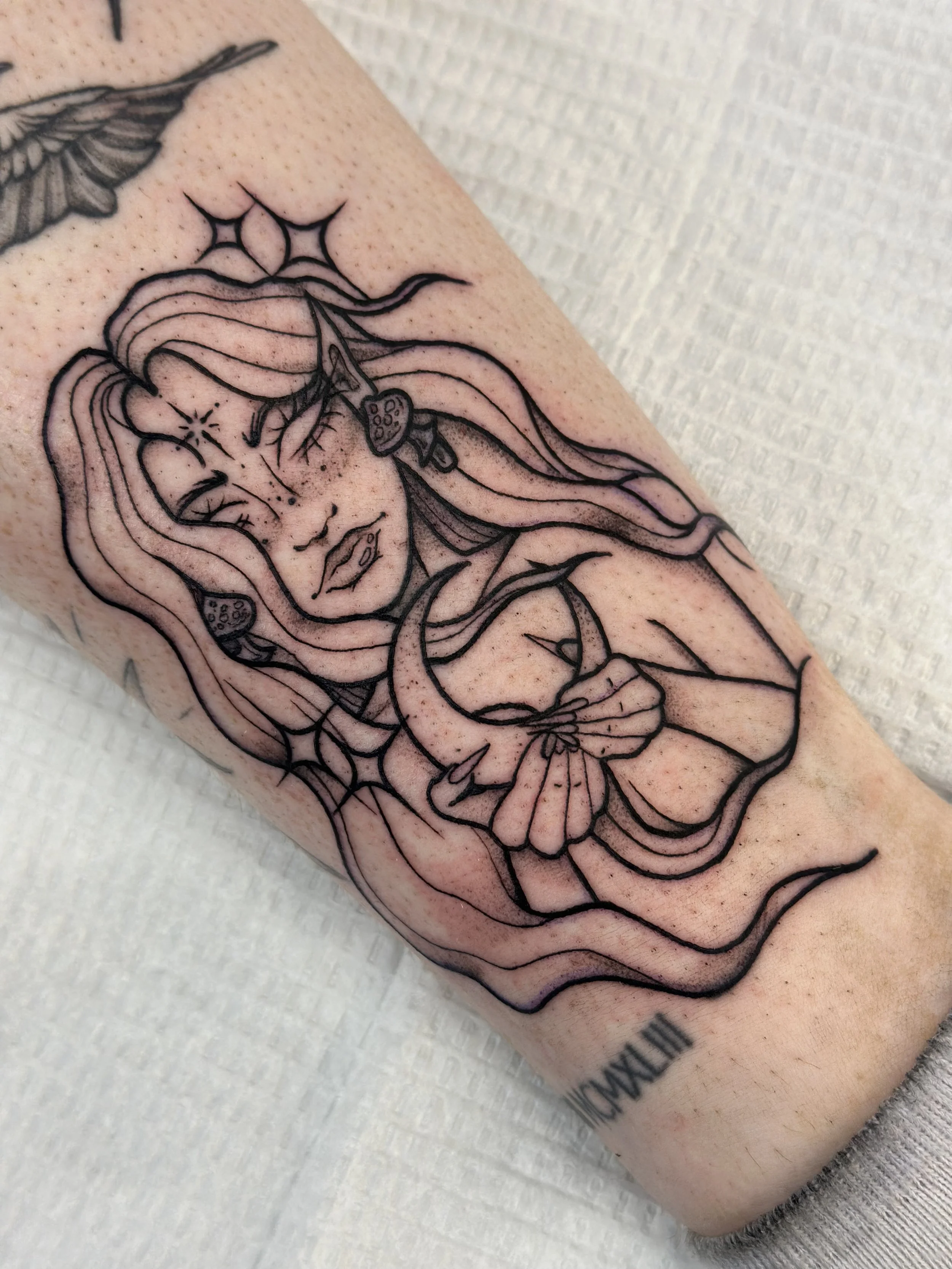 Tattoo of a woman with closed eyes, long flowing hair, surrounded by cobwebs, holding a crescent moon with a bow, and with a fairy-like appearance.
