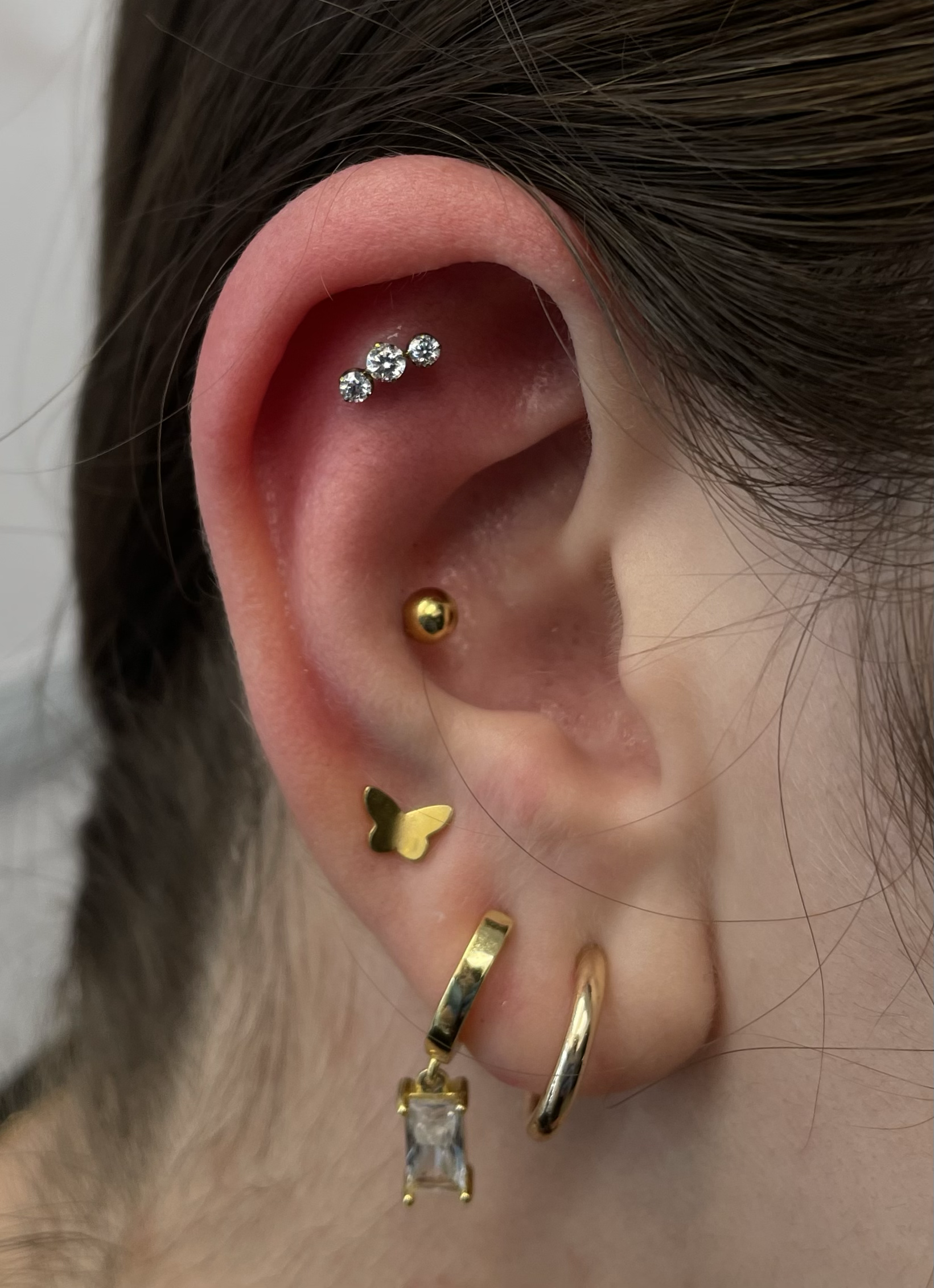 Close-up of a person's ear with multiple piercings, including a cartilage piercing with three small diamonds, a helix piercing with a gold ball, a lobe piercing with a gold butterfly-shaped stud, a hoop earring with a clear rectangular gemstone, and 