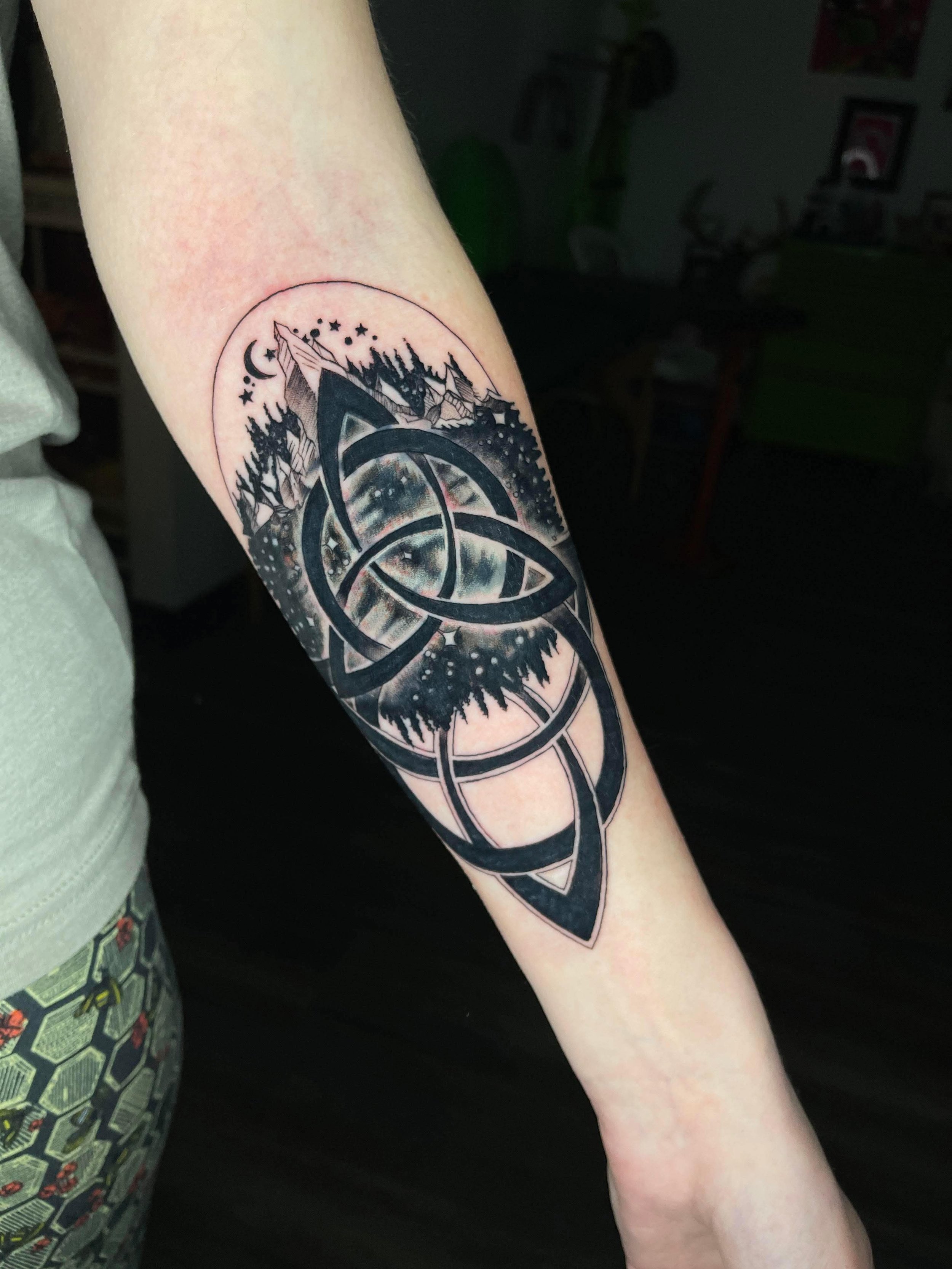 A black and gray tattoo featuring intricate Celtic knotwork, a forest scene with mountains, trees, and a night sky with stars, moon, and an eye in the background on a person's forearm.