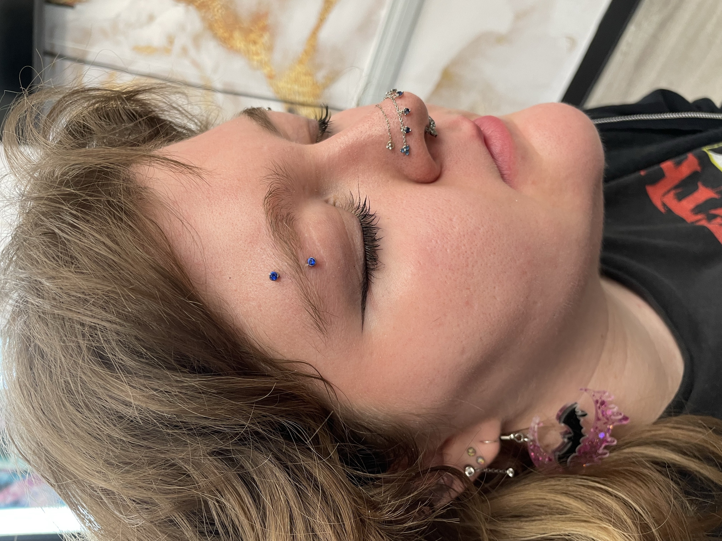 Close-up of a woman with various facial piercings, including a septum piercing, eyebrow piercings, and earrings, with makeup and styled hair, lying with her eyes closed.