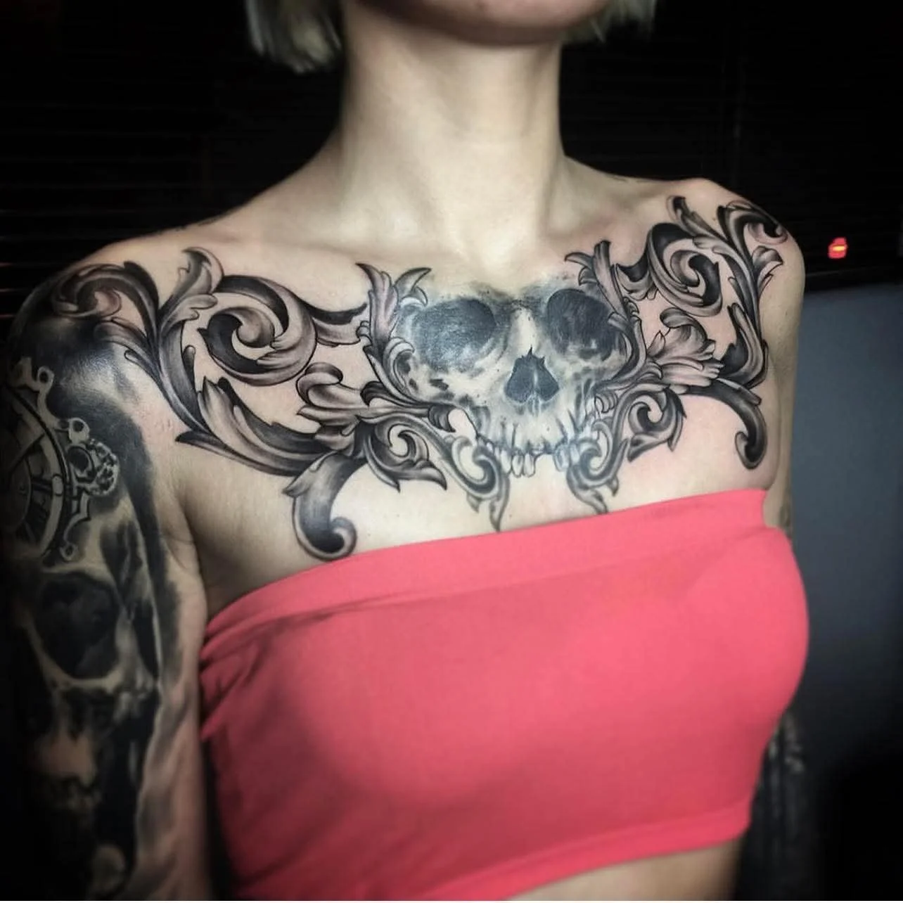 A woman wearing a pink tube top showcases a large tattoo across her chest and shoulders, featuring a detailed skull with floral and ornamental elements in black and gray ink.
