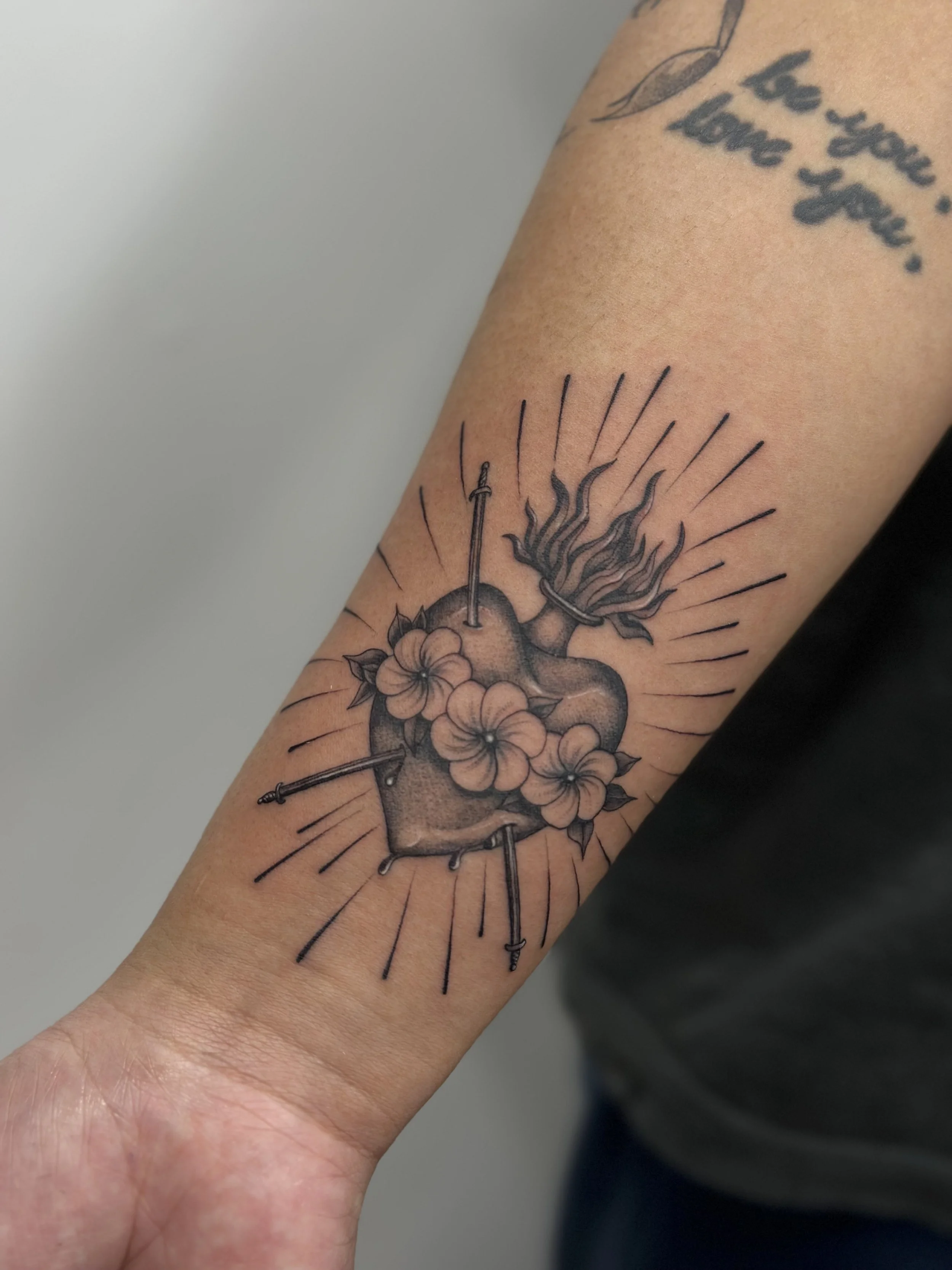 Tattoo of a heart with flowers, surrounded by radiating lines, with a sword piercing the heart from above and flames coming out of the top.