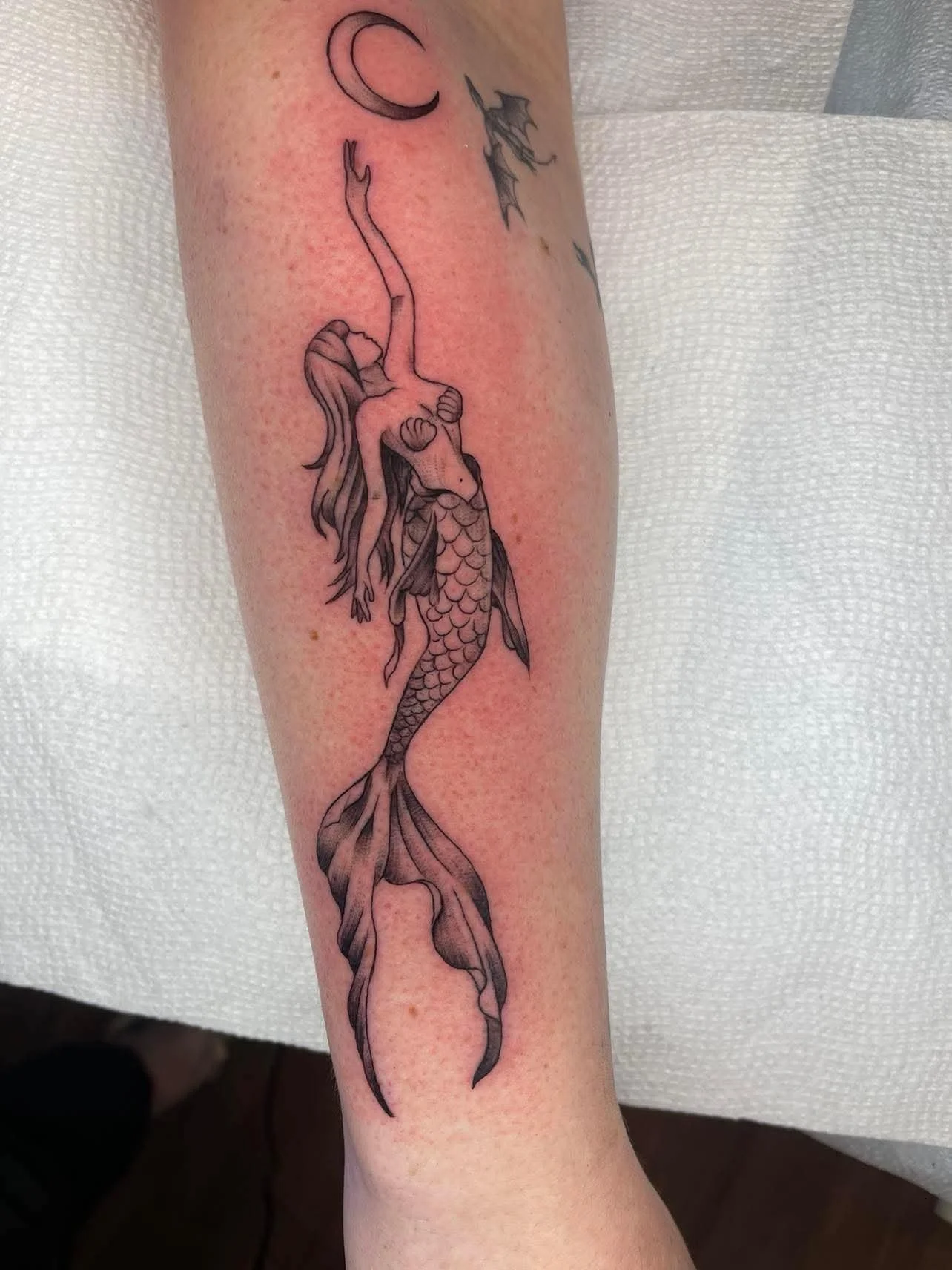 Black and gray tattoo of a mermaid with a fish tail on a person's arm, with a moon and a dragonfly nearby.