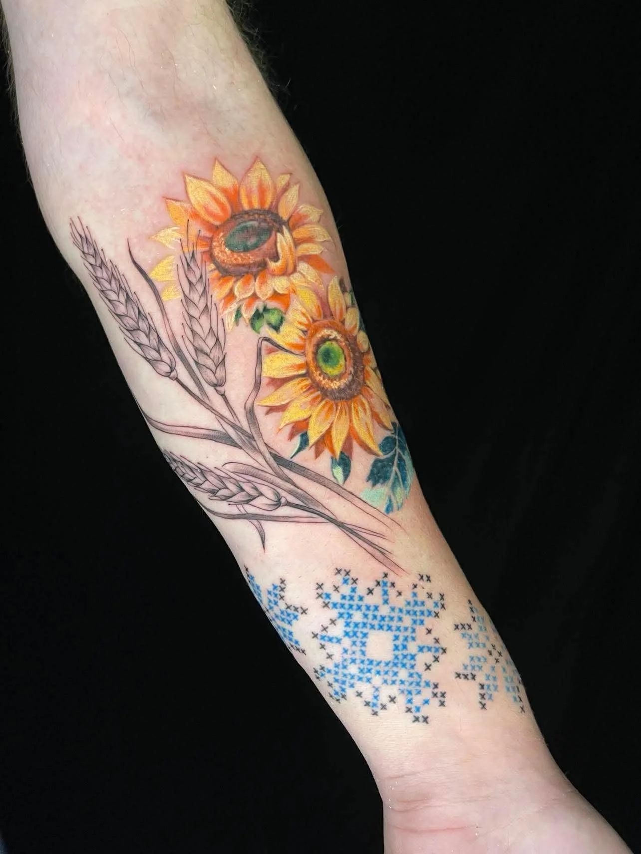 Tattoo of two sunflowers and wheat stalks on a person's forearm, with a design resembling a pixelated snowflake below the flowers.