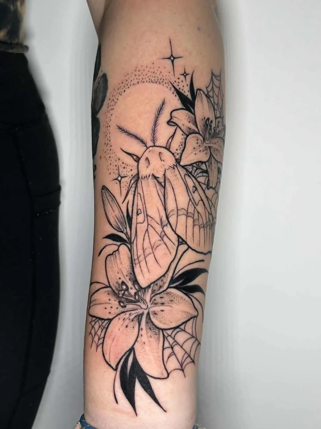 A black and gray line art tattoo on the arm featuring a moth with detailed wings, surrounded by lilies, with a dotted circle and sparkles in the background.