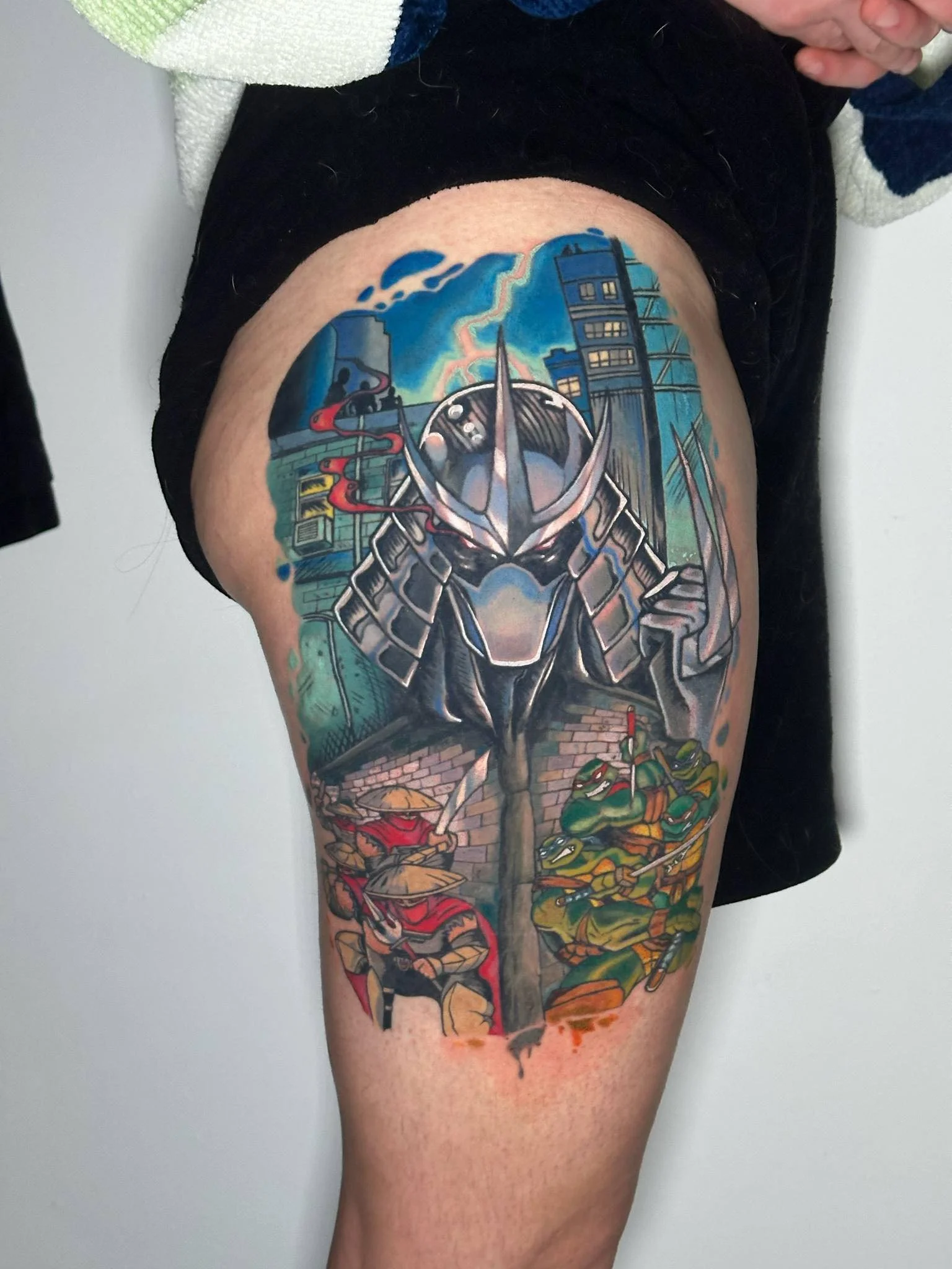 A detailed colorful tattoo on a person's thigh featuring a futuristic robot or warrior in the center, with a cityscape and lightning in the background, and ninja-like characters with swords and masks at the bottom.