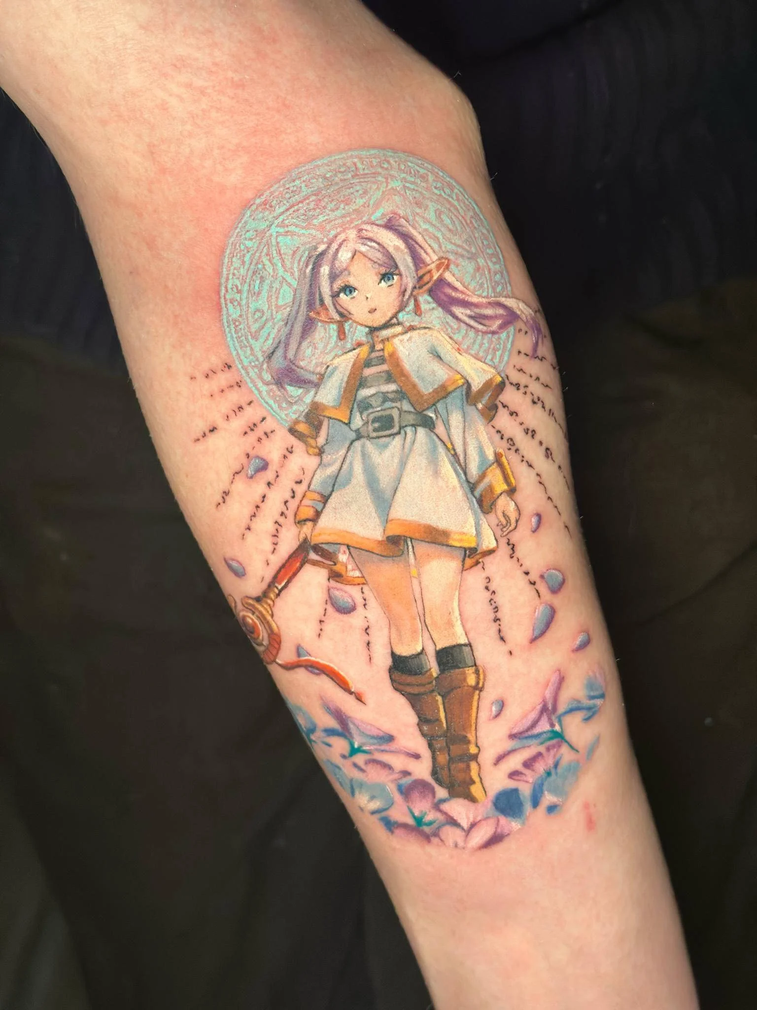 Colorful tattoo of a fantasy female elf character with purple hair, blue eyes, and pointy ears, holding a staff, surrounded by a circular magical symbol and decorative pink and blue elements.