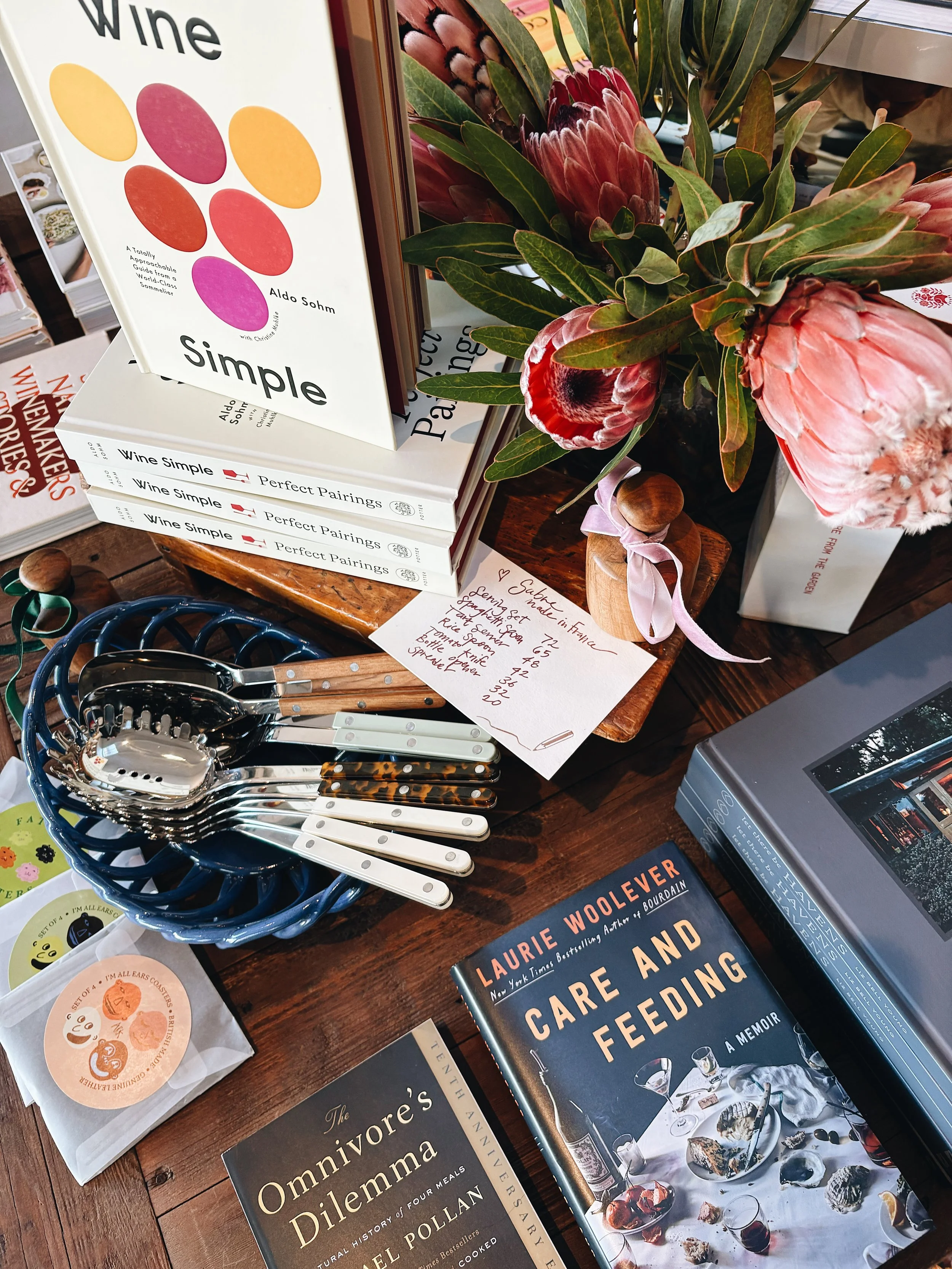 A table displays a stack of books, a bouquet of flowers, a selection of silverware, and a handwritten note with a wine list, all arranged for a book signing or event.