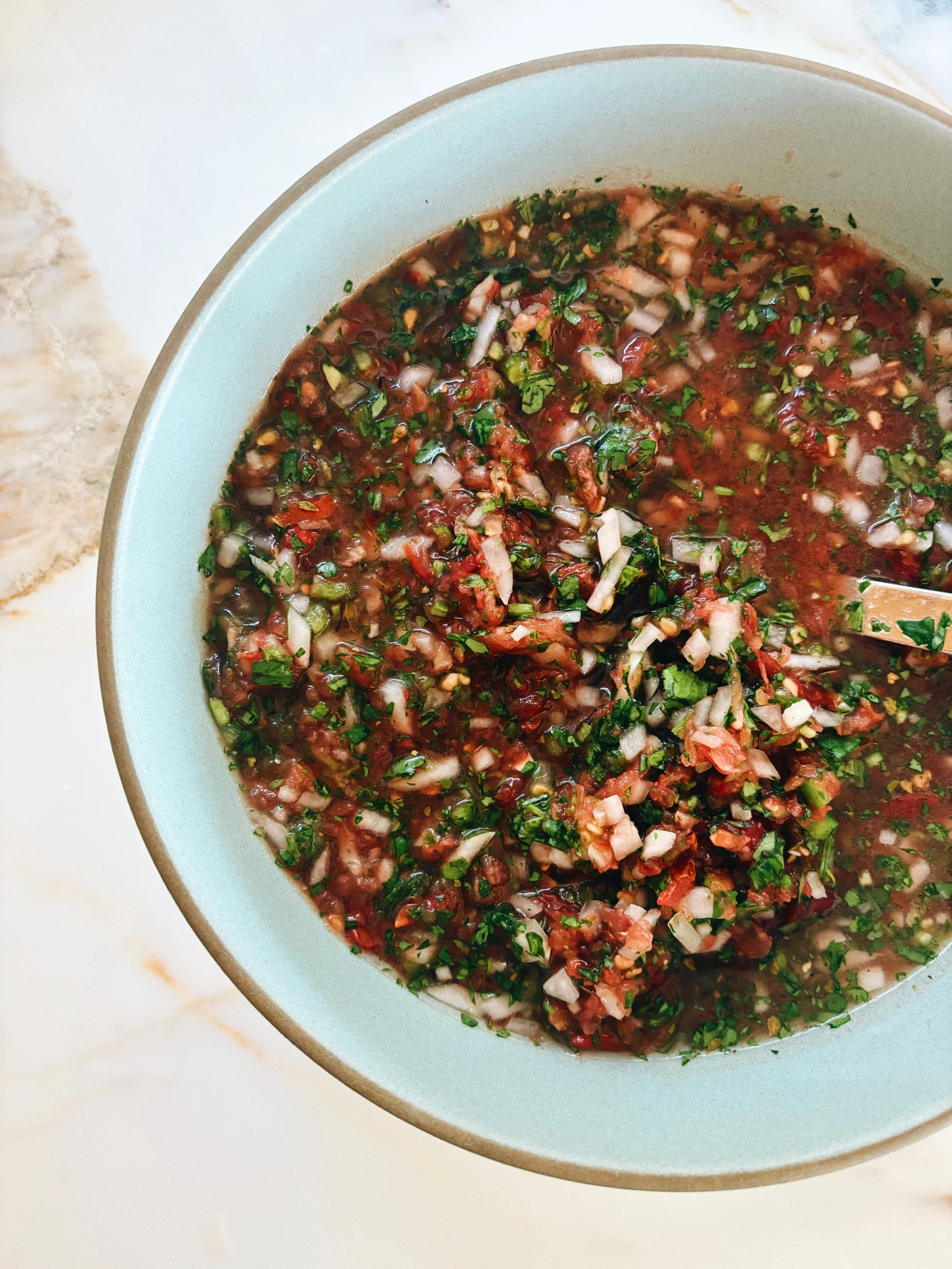 A bowl of pico de gallo salsa with chopped tomatoes, onions, cilantro, and green peppers on a light-colored marble surface.