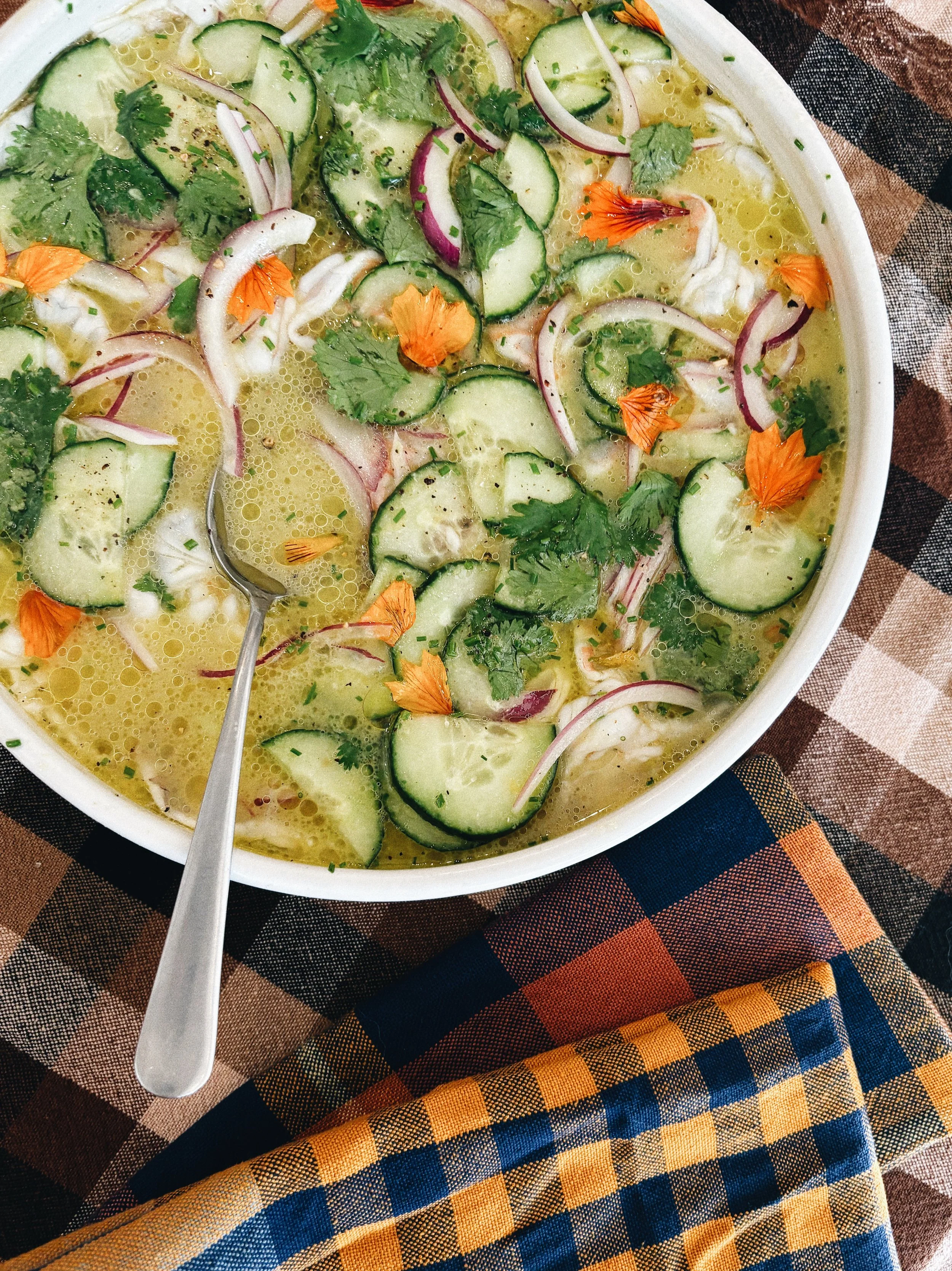A bowl of chicken lime soup garnished with cucumber slices, red onions, cilantro, and edible flower petals, with a spoon resting inside, on a checkered tablecloth with colorful napkin.