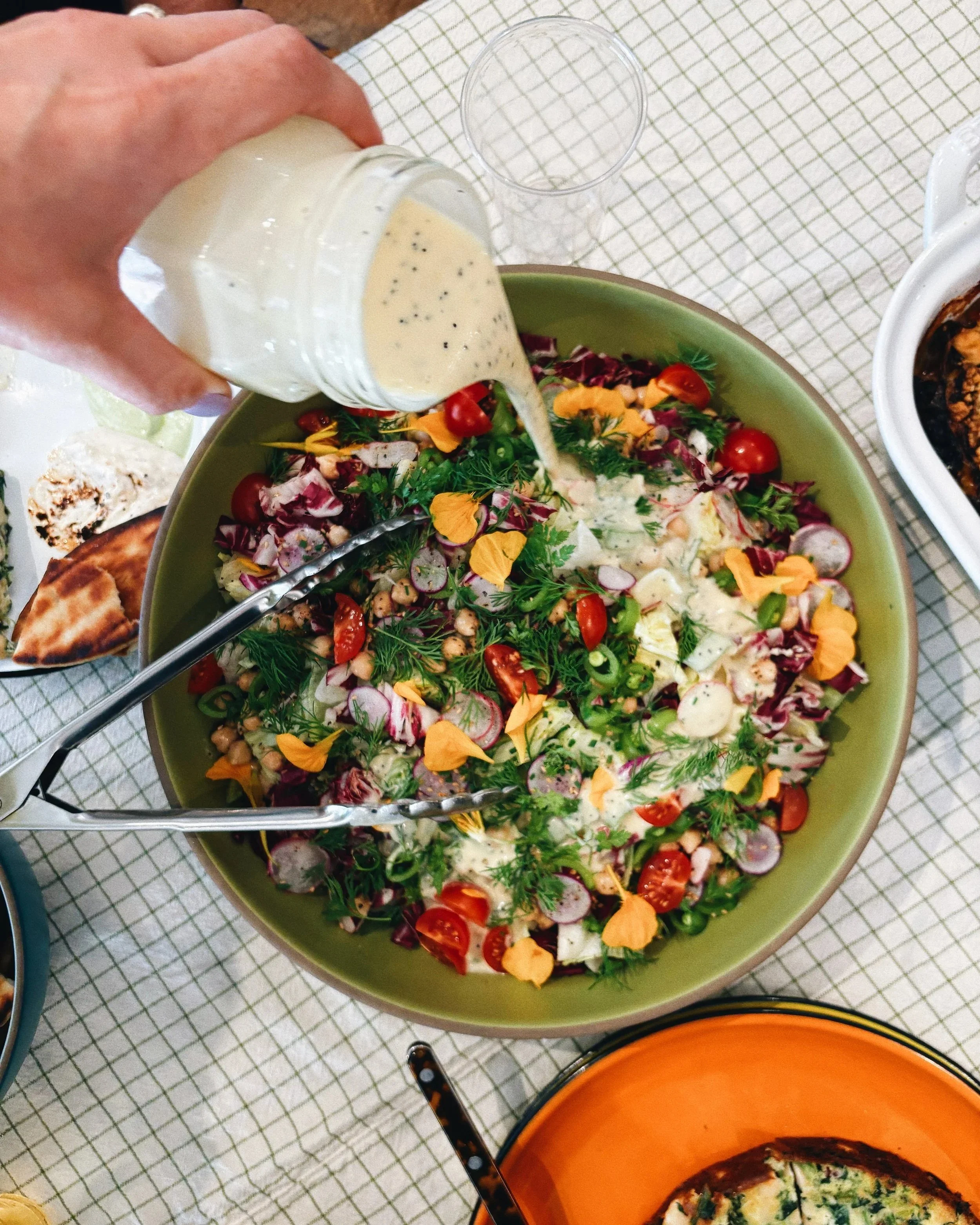 A large bowl of mixed vegetable salad with cherry tomatoes, radishes, greens, and edible flower petals being topped with creamy dressing.