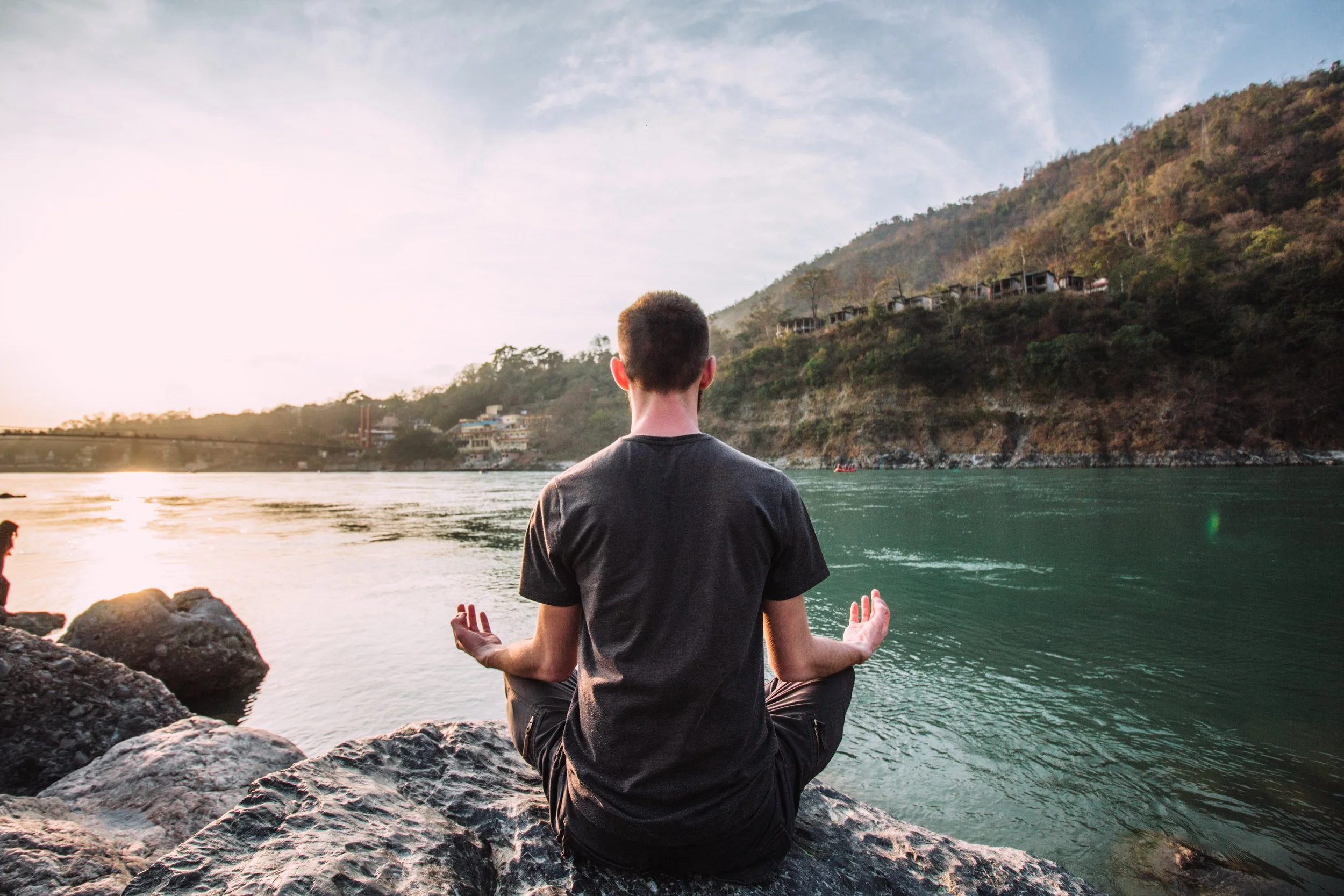 A man practicing yoga or meditating on a rock by a river at sunset, with mountains and houses in the background.