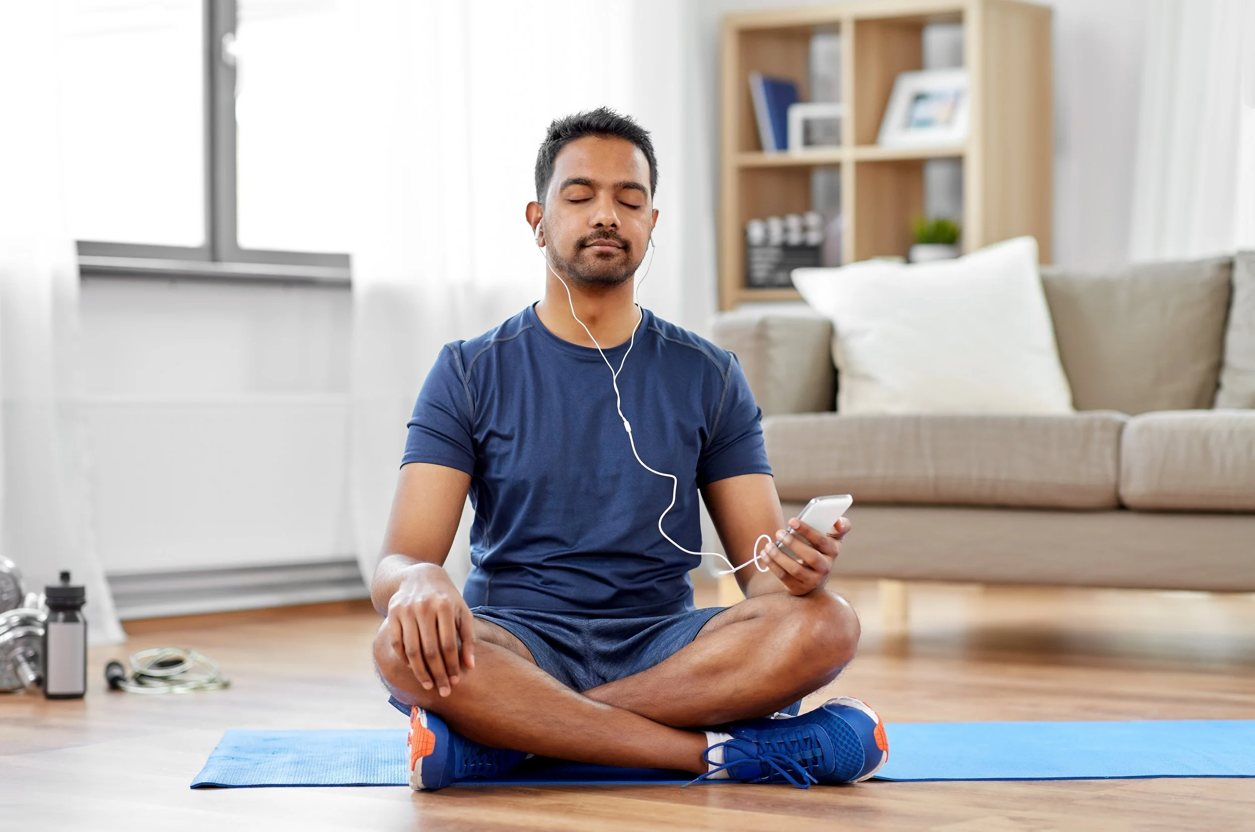 A man sitting cross-legged on a yoga mat in a living room, wearing workout clothes and listening to music through earphones while holding a smartphone.