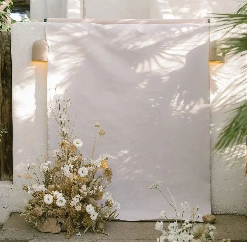 A white fabric backdrop with shadows from sunlight and leaves, decorated with a bouquet of dried white, beige, and brown flowers and rocks at the base.