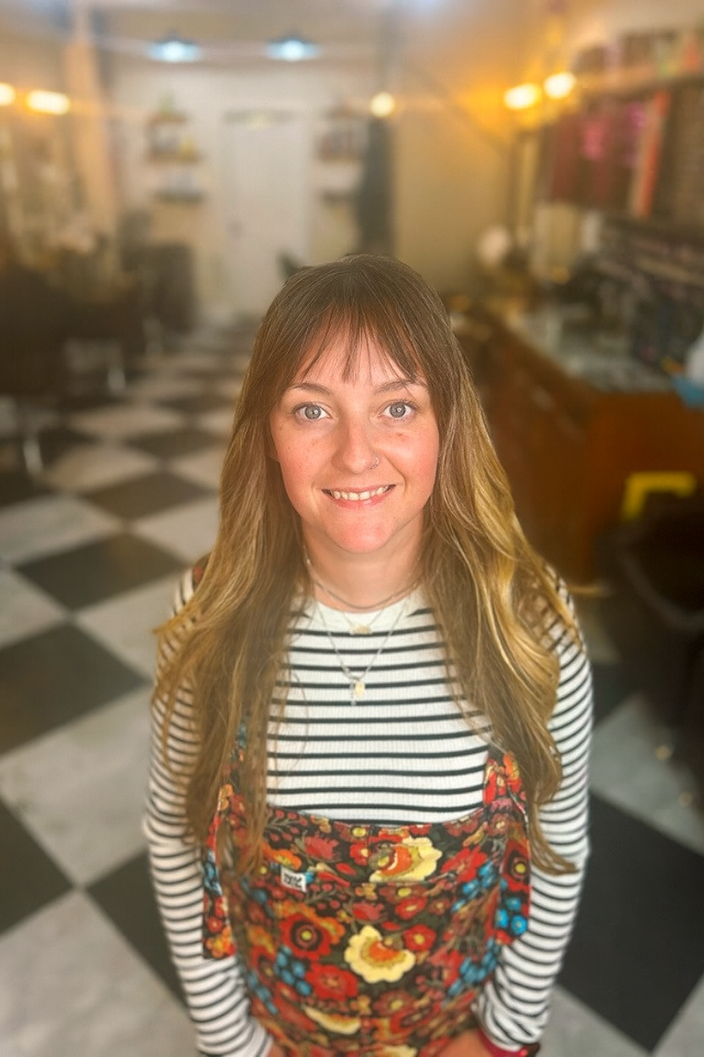 A young woman with shoulder-length brown hair, striped shirt, and floral apron standing in a hair salon with a checkered floor and blurry salon background.