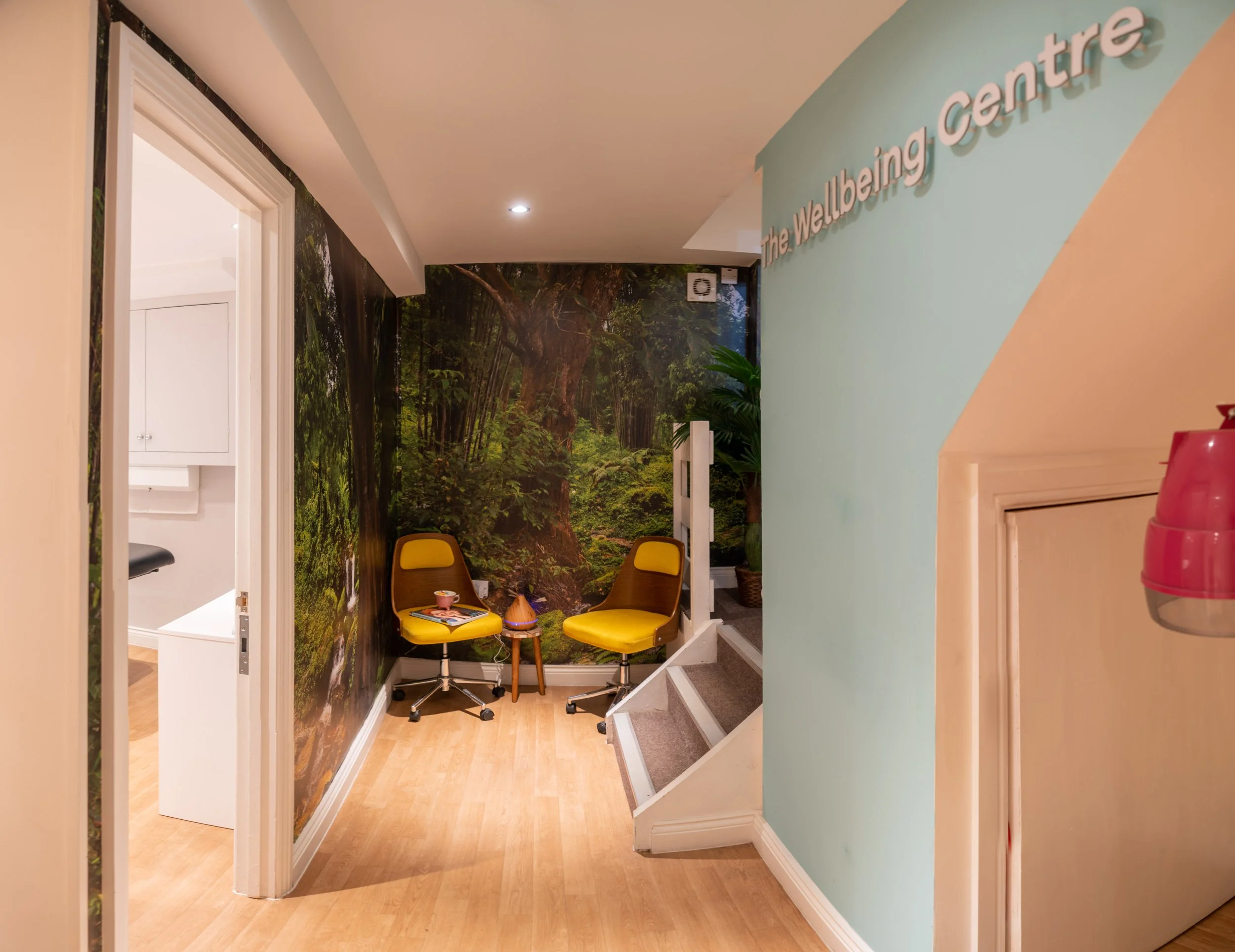 Waiting area with two yellow chairs, a small table with a teacup, and a forest mural wall at The Wellbeing Centre.