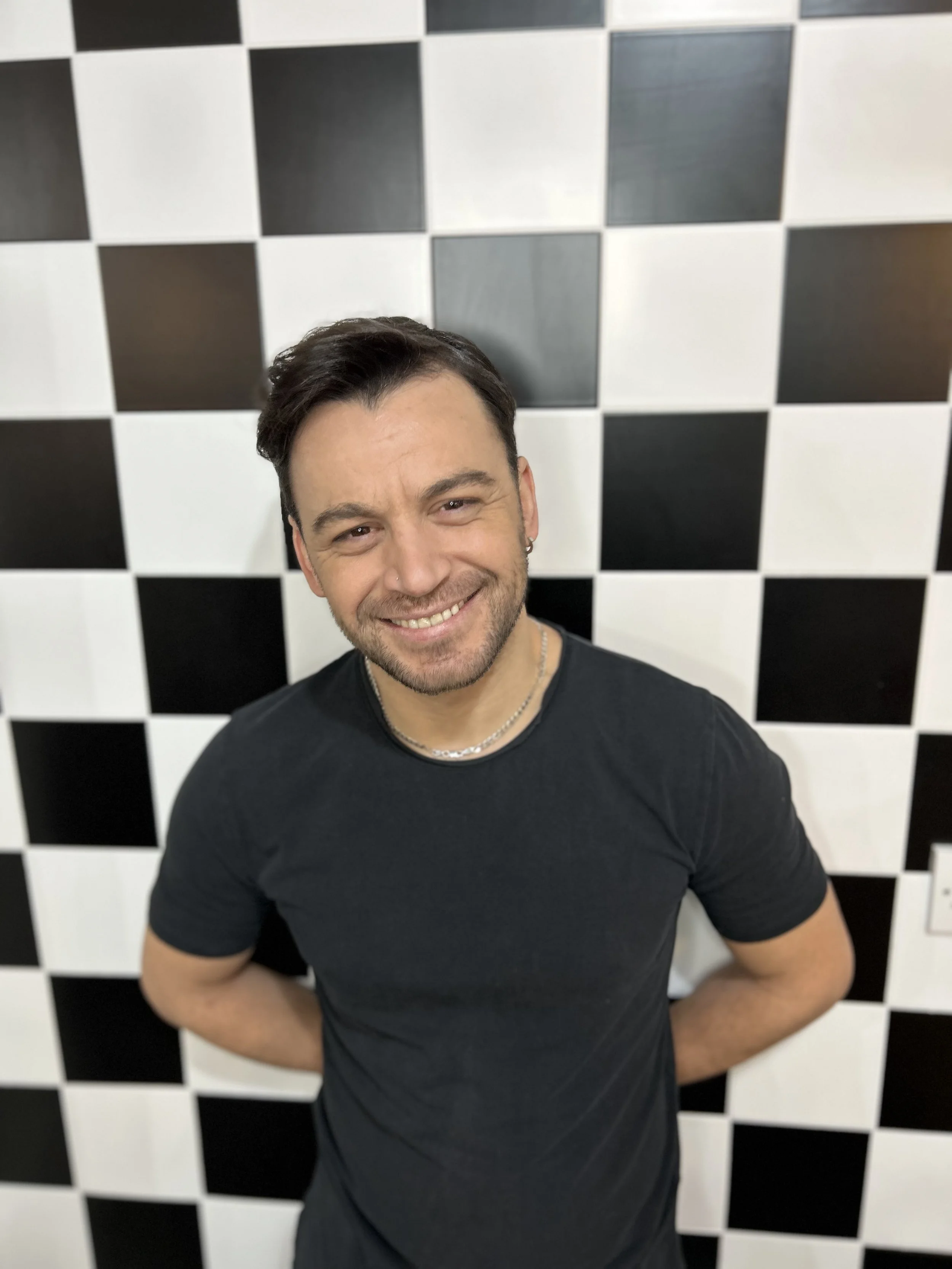 A smiling man with dark hair, wearing a black t-shirt, standing in front of a black and white checkered wall.