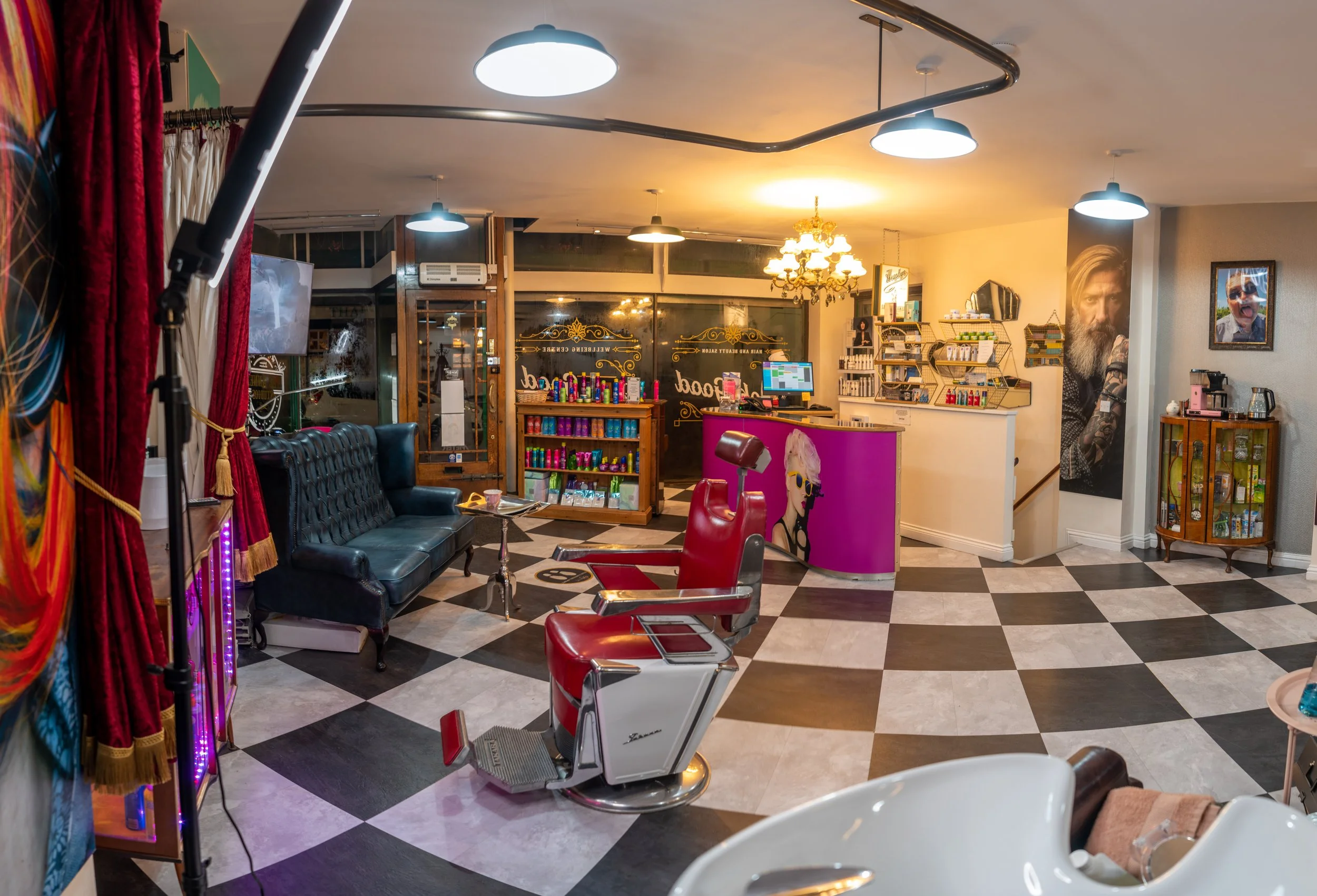Interior of a vintage-style barber shop or salon with checkered black and white floors, red barber chairs, a black couch, a reception desk with colorful decorations, and various shelves and framed photographs on the walls.