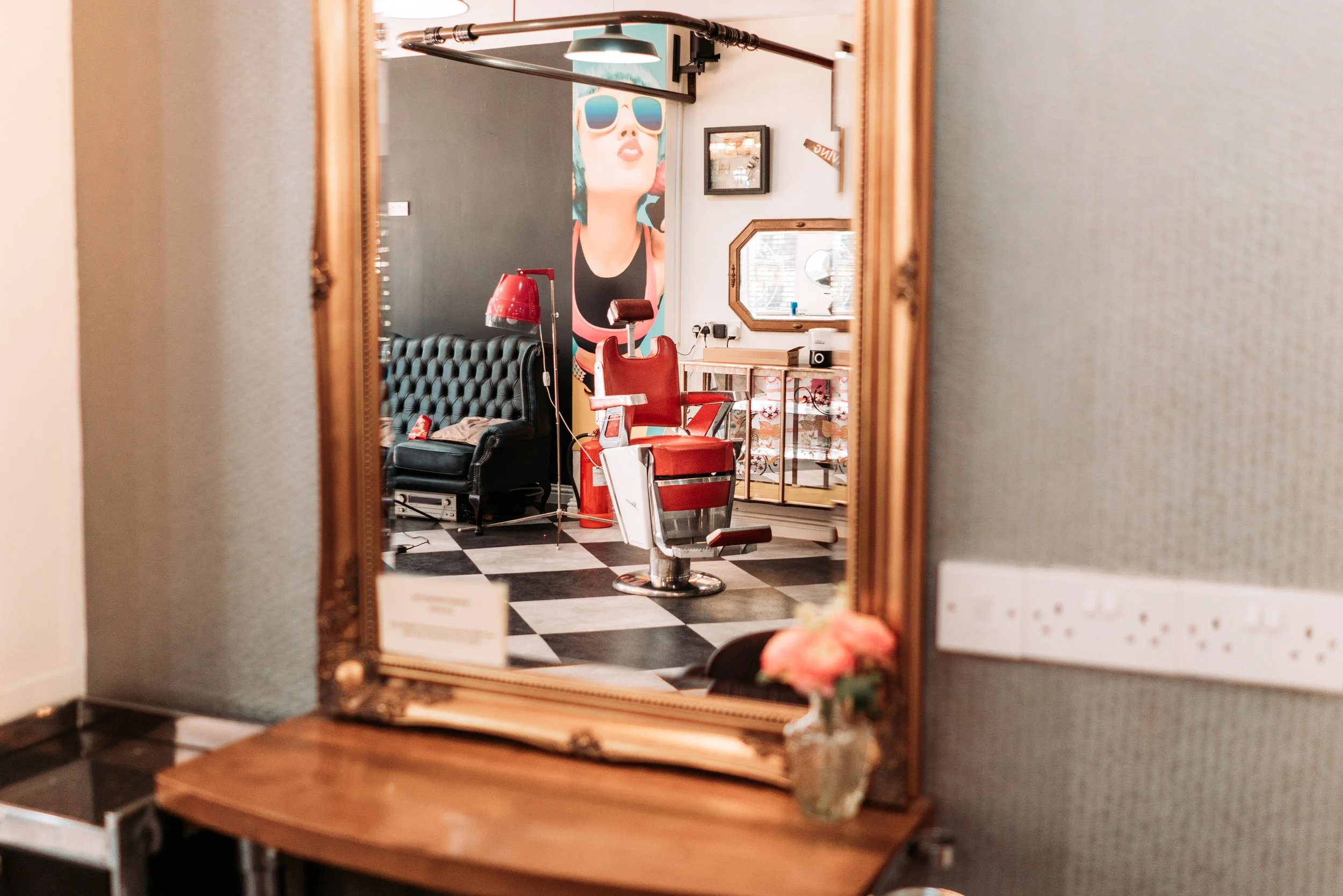 A vintage barber or salon chair with red cushioning, reflected in a decorative gold-framed mirror, with a black tufted sofa, a red floor lamp, and a large wall art of a woman with sunglasses in the background.