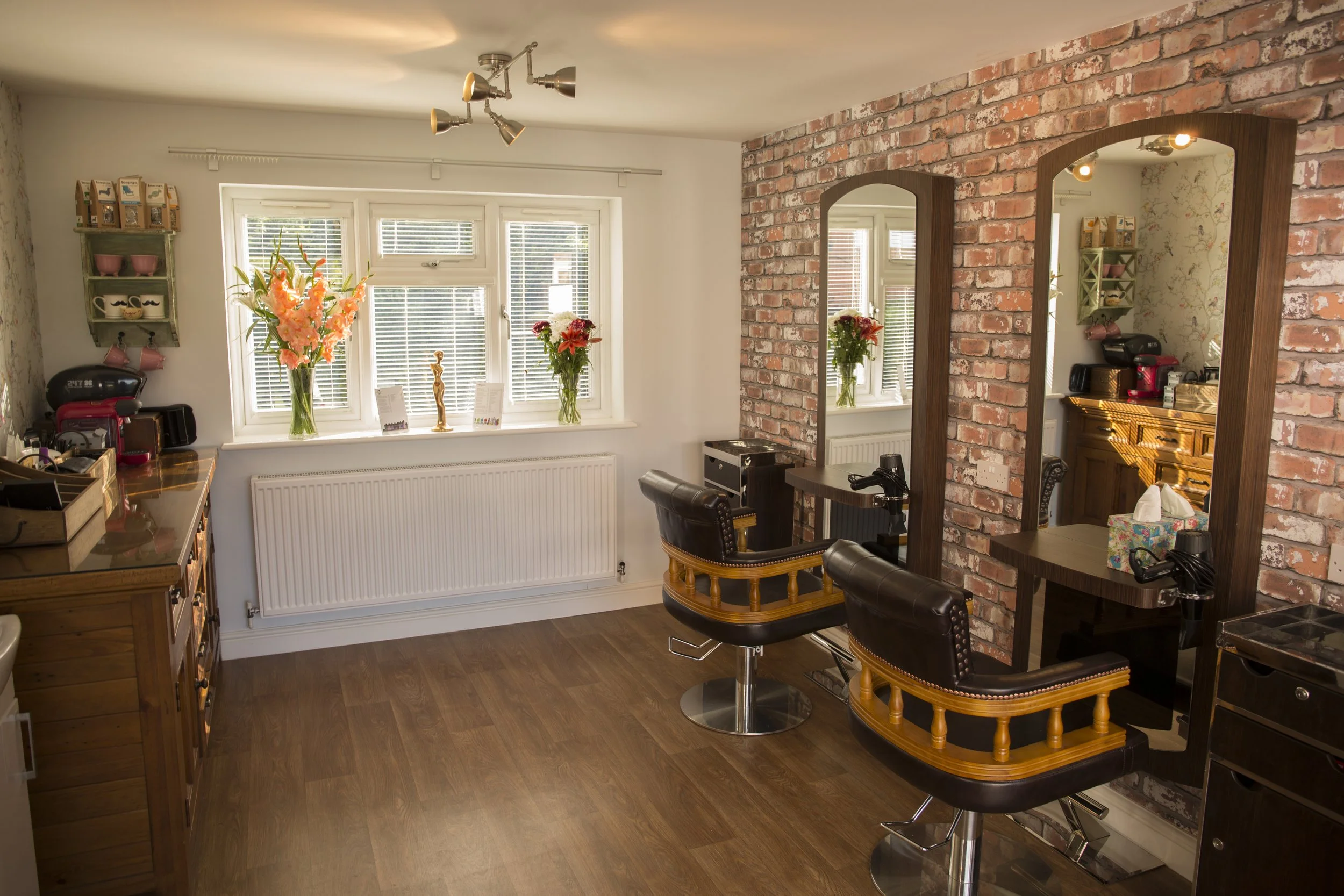 Interior of a hair salon with two styling stations featuring large mirrors, wooden furniture, and salon chairs, brick accent wall, wooden floors, and a window with flowers on the window sill.