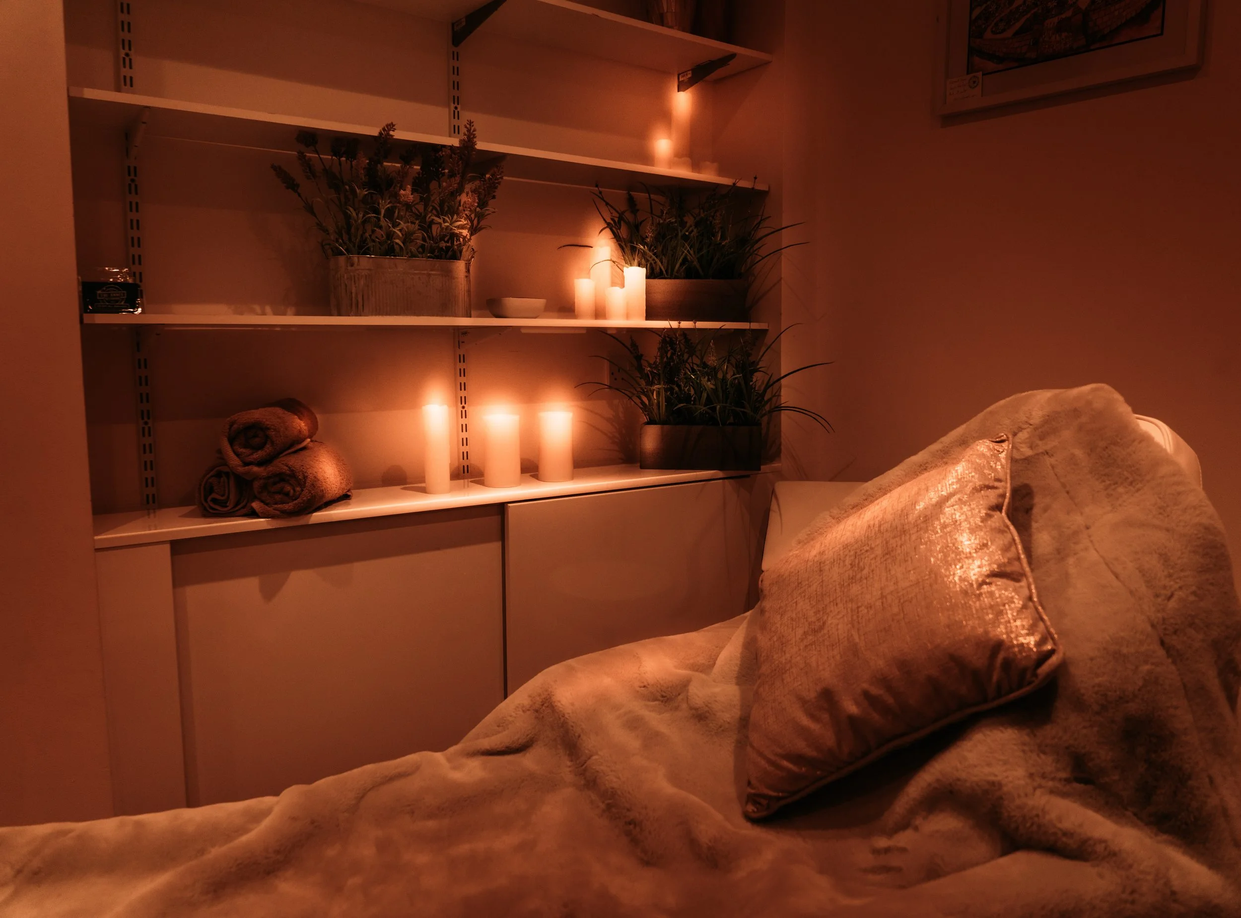 A cozy room with dim lighting, featuring three candles on a white shelf, two potted plants, and rolled towels. A plush pillow sits on a beige sofa or bed in the foreground.