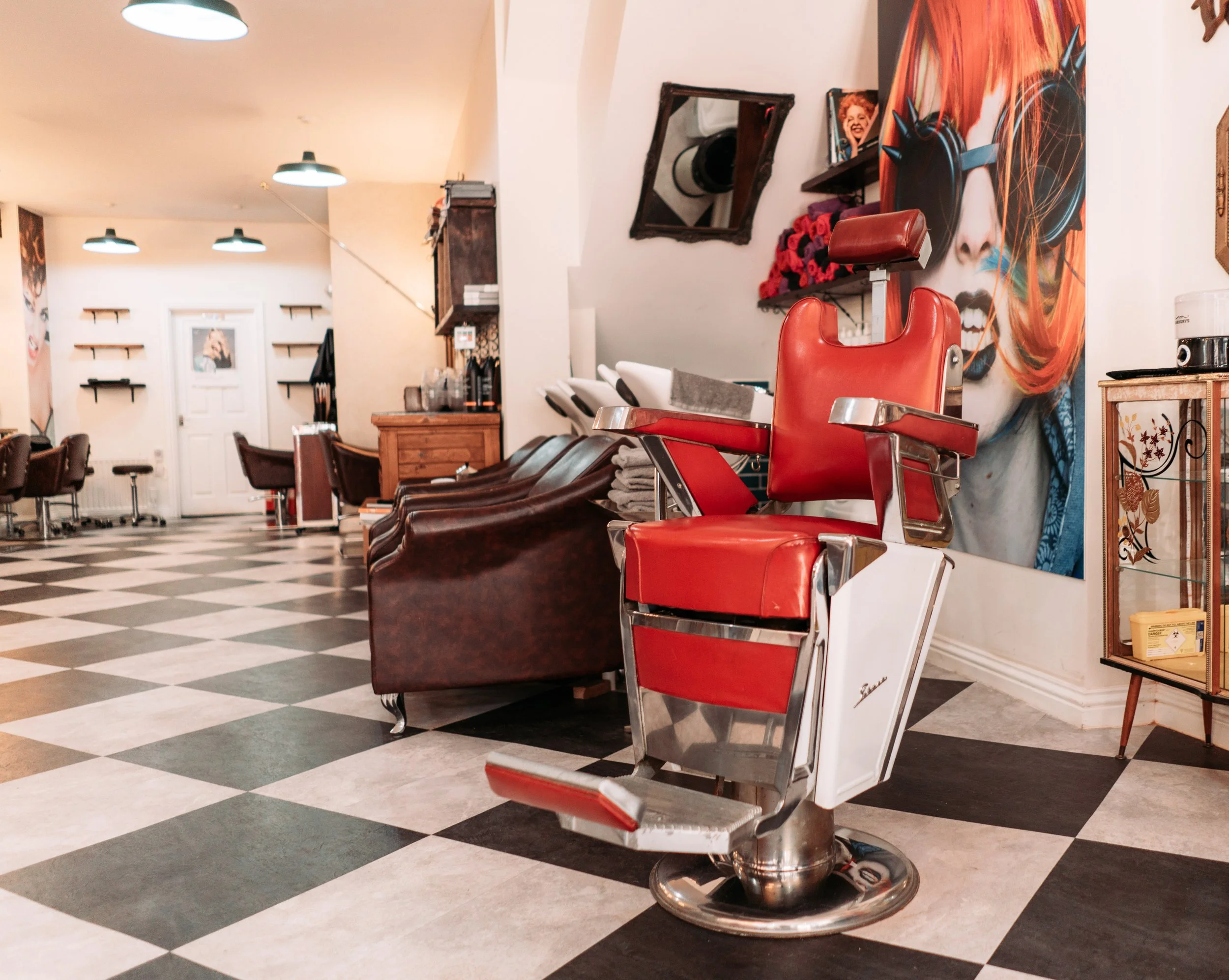 Red vintage barber's chair in a hair salon with checkered black and beige floor, artwork on the walls, and styling chairs in the background.