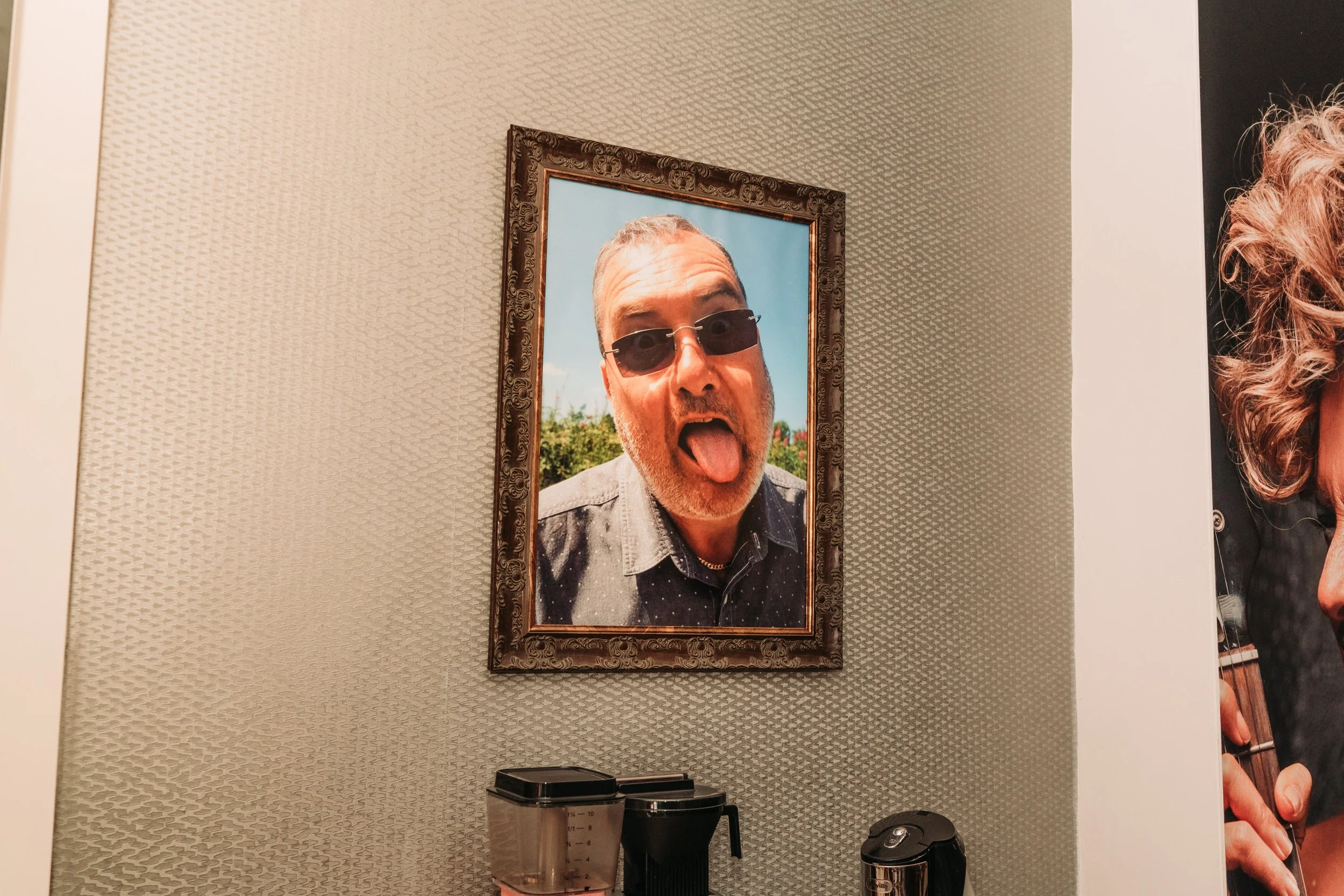 A framed photo of a man with sunglasses, sticking his tongue out, hanging on a textured wall.