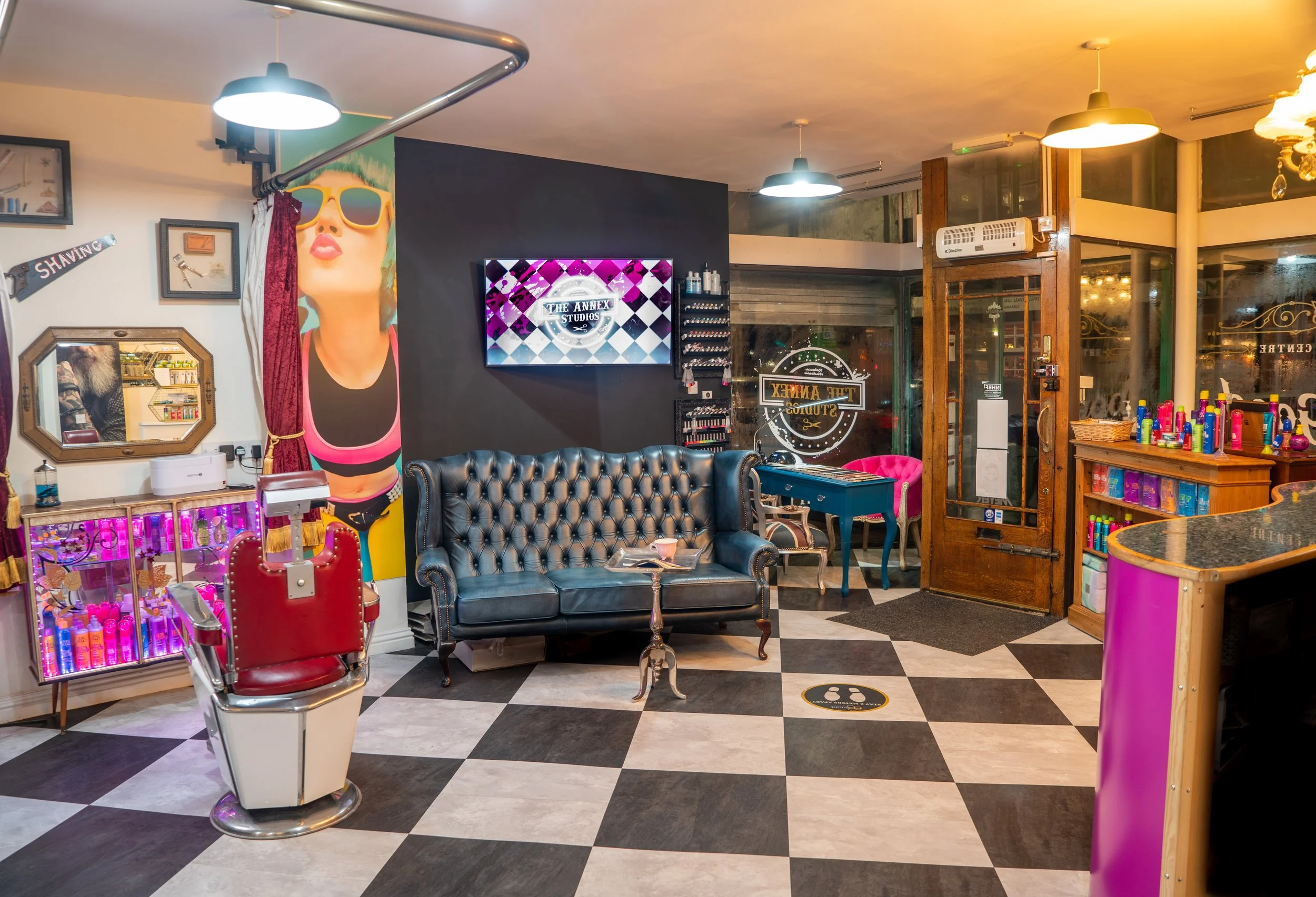 Interior of a beauty salon with vintage-style decor, featuring a black tufted sofa, a small round table, a pink chair, shelves with hair products, and colorful artwork on the walls.