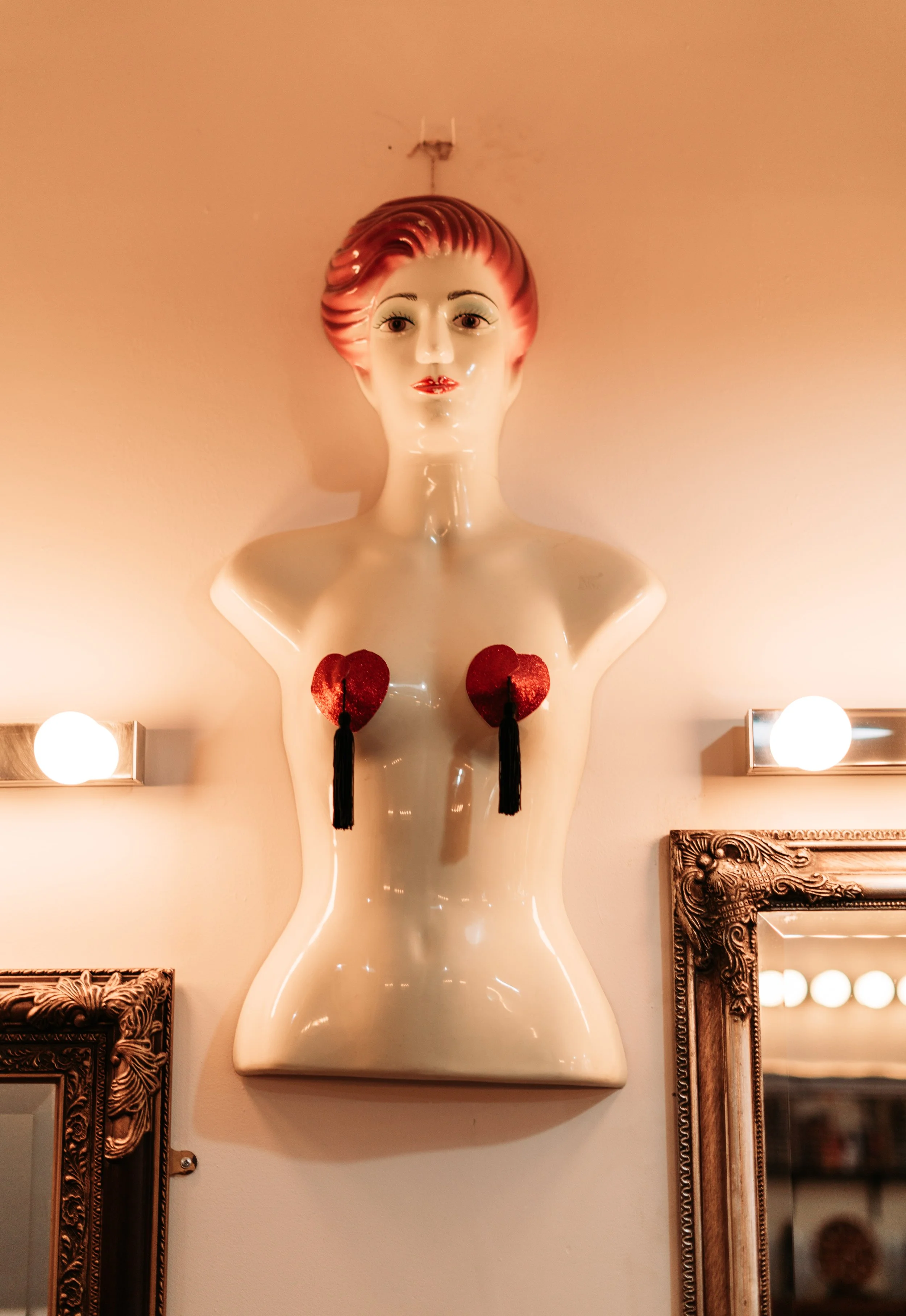 Decorative wall sculpture of a stylized female bust with a retro hairstyle, red heart-shaped pasties with black tassels, and bright lighting fixtures on either side.