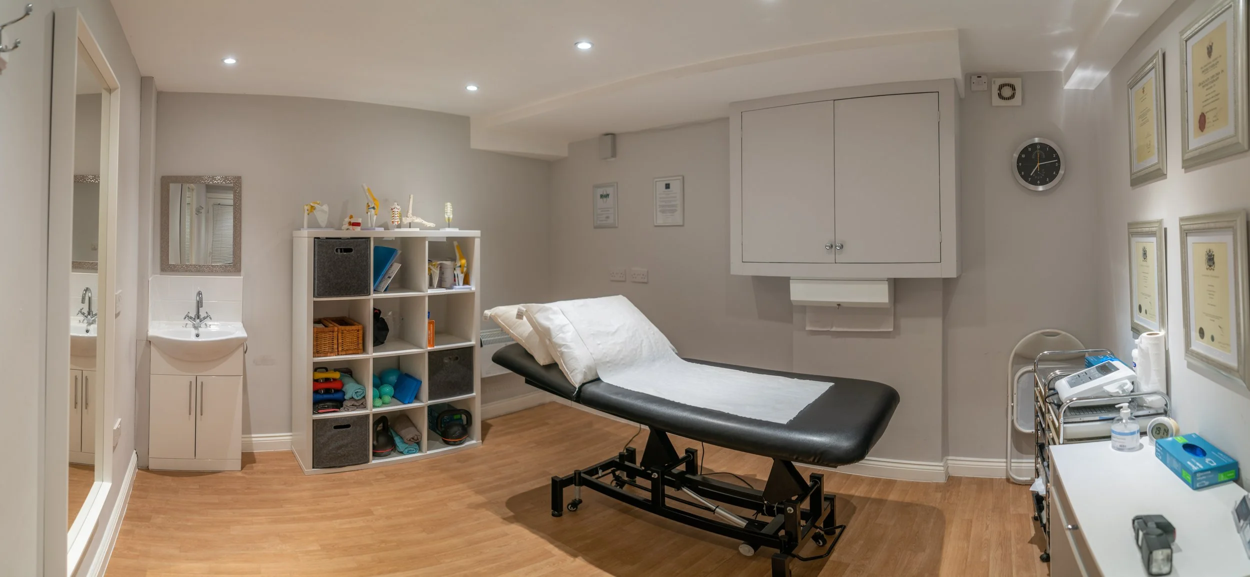 Medical examination room with an examination table, storage shelves with medical supplies, wall-mounted certificates, a clock, a mirror, a sink, and various medical equipment.