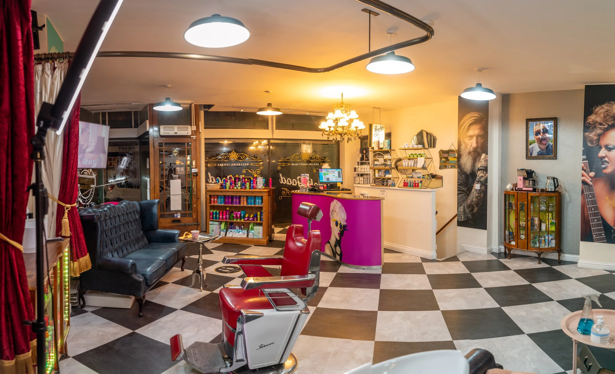 Interior of a modern hair salon with a black leather sofa, a vintage barber chair in red and white, a checkered black and white floor, a reception desk with a vibrant pink front featuring a drawing of a woman with blonde hair and sunglasses, various hair products on shelves behind the desk, large posters of people with styled hair on the walls, and decorative lighting including ceiling lamps and a chandelier.