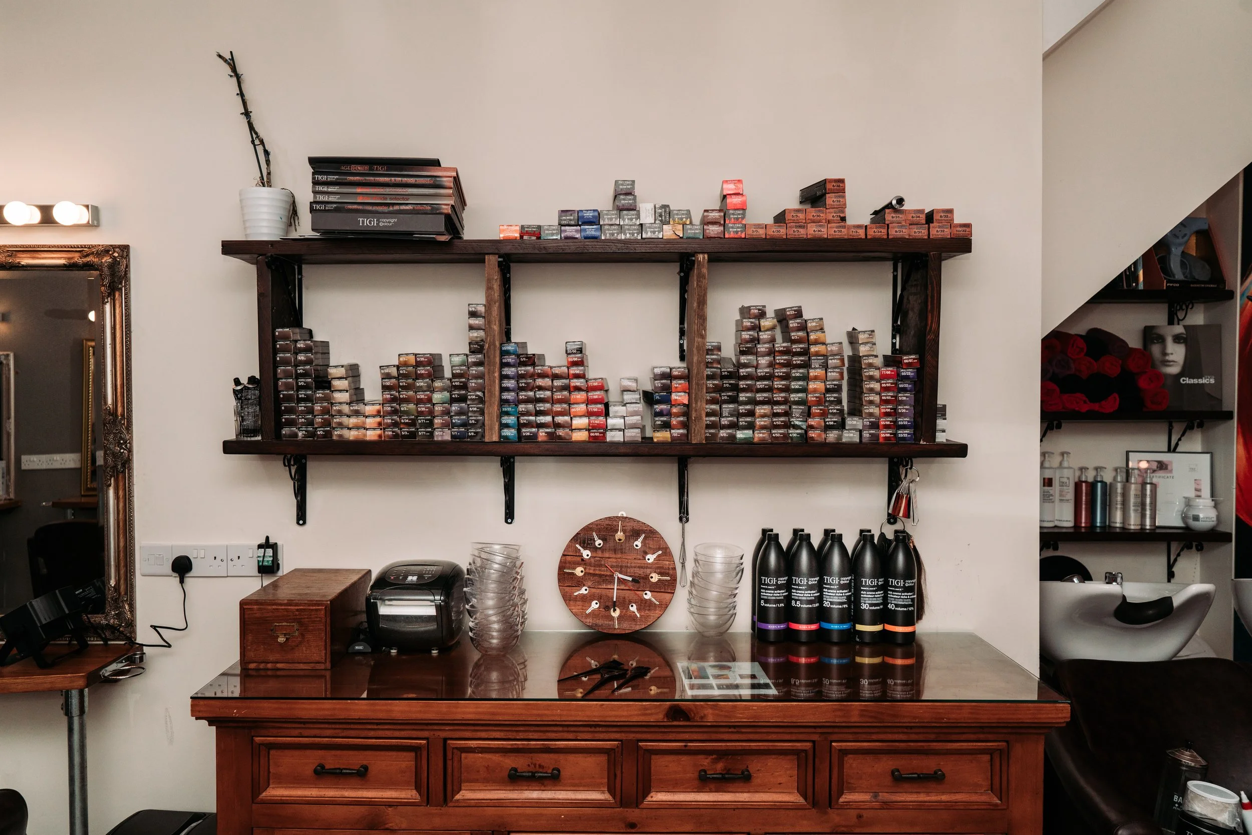 Wooden shelf with various hair dye boxes, a white vase with a branch, a clock, and hair products on a wooden countertop in a salon.
