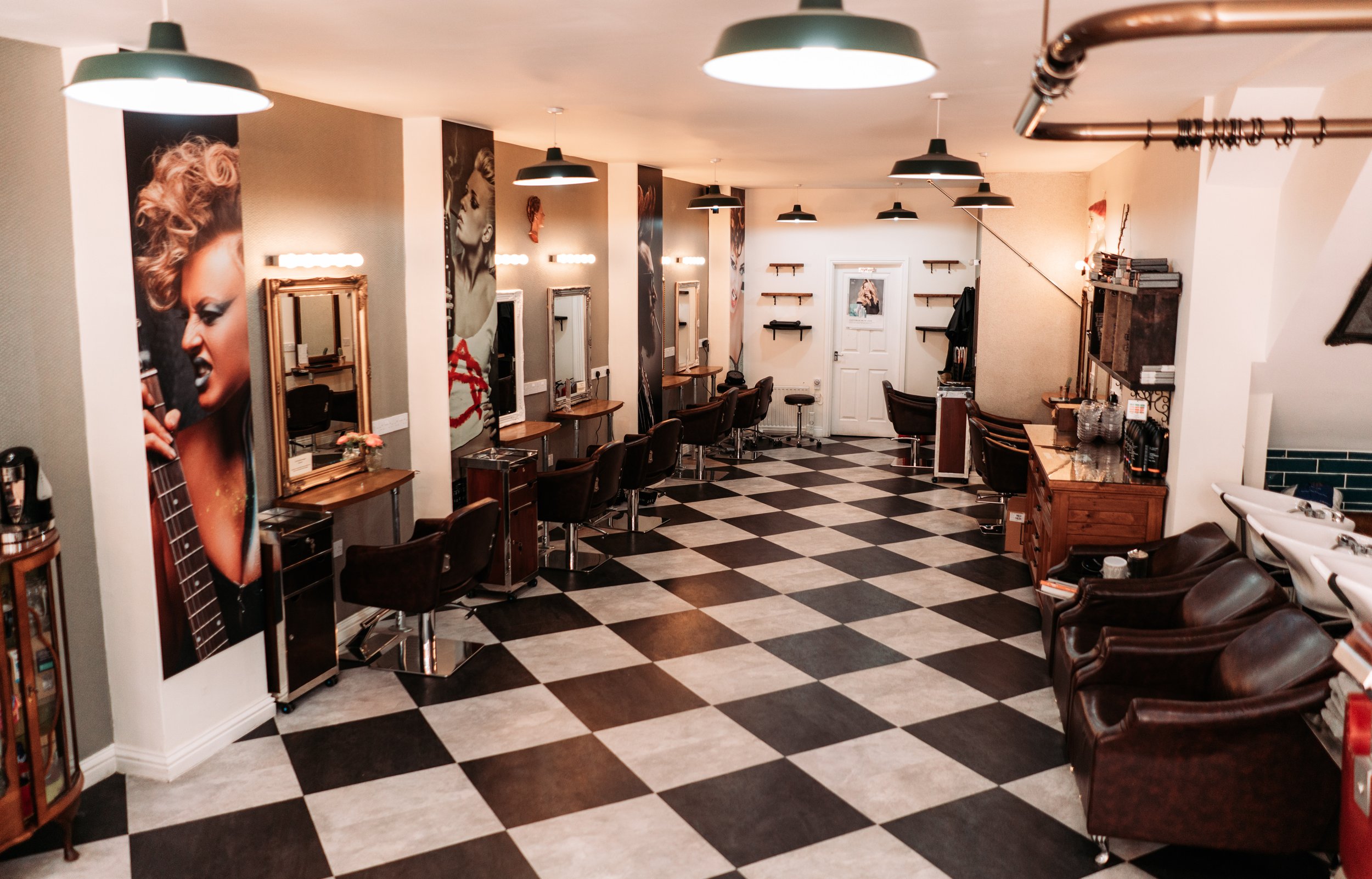 Empty hair salon with multiple styling stations, mirrors, and chairs, some with hairstyles displayed on the walls, checkered black and white floor, and salon furniture.